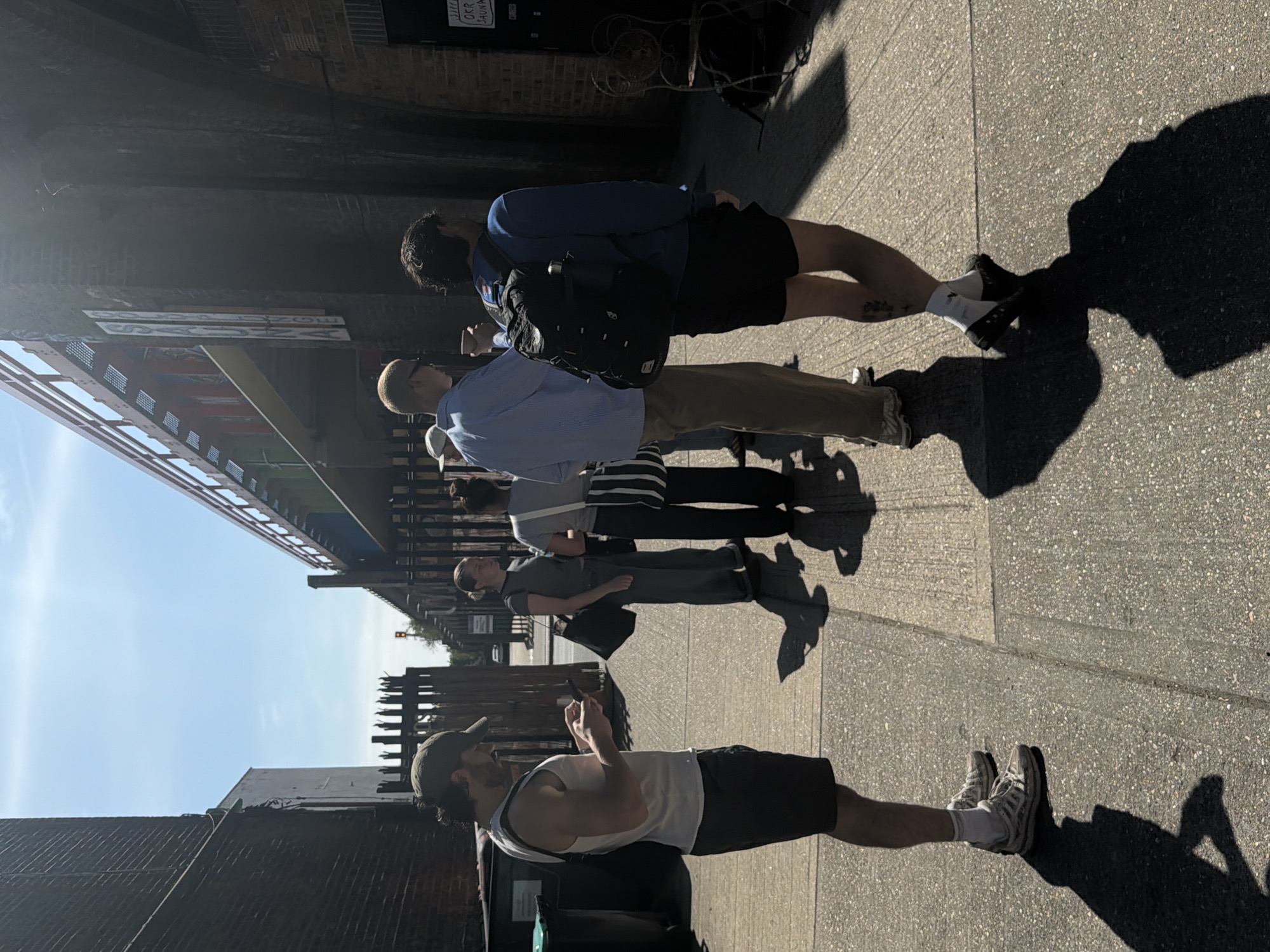 A group of five people stands on a sidewalk under a metal structure, likely a railway bridge, on a sunny day. The people are walking towards the right. The first person is wearing a white tank top, dark shorts, and a baseball cap. The next person is wearing a grey t-shirt and gray pants, carrying a black purse. The third person is wearing a grey shirt and black and white striped shorts. The fourth person is wearing a light blue shirt and khaki pants. The final person is wearing a blue sweatshirt and black shorts, and a black backpack. Shadows from the people and the metal structure are cast on the sidewalk. The location is possibly in London, United Kingdom.