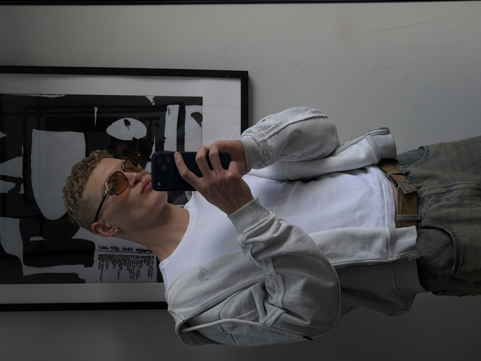 A young man with blonde curly hair and sunglasses is taking a selfie in a room. He is wearing a white t-shirt, a grey zip-up hoodie, and blue jeans. He is holding a dark-colored smartphone in his right hand. Behind him, there is a framed black and white abstract artwork hanging on the wall, and to the right of the artwork is a plain white wall. The room appears to be indoors, and there are no visible indications of the specific location within London, United Kingdom.