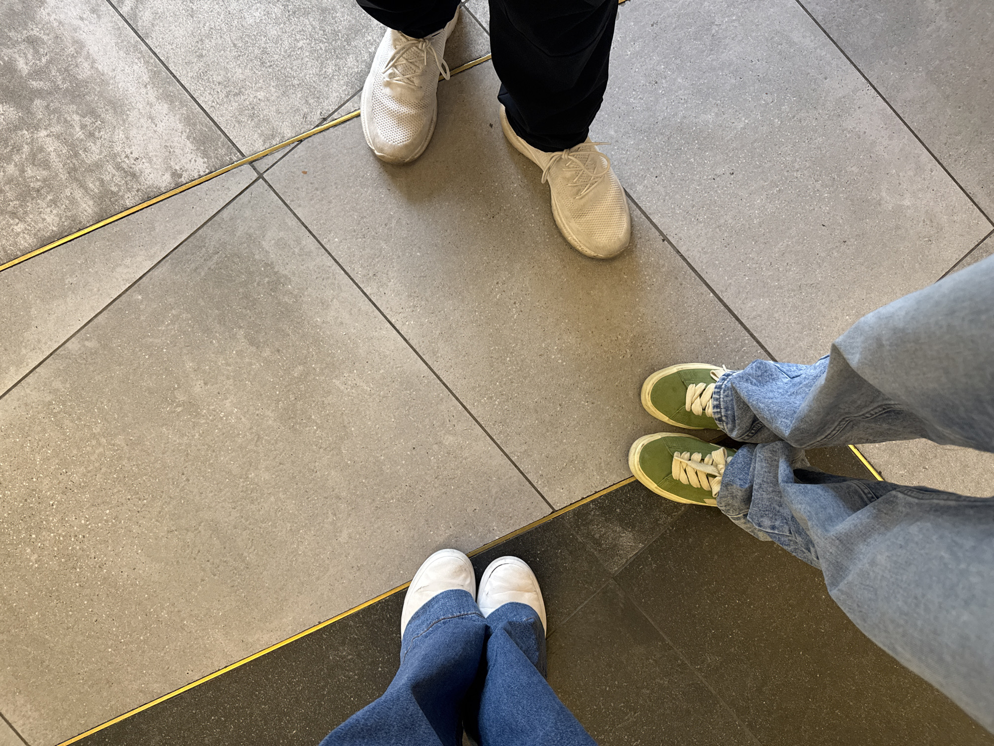 The image depicts a section of flooring with four pairs of legs and feet. The flooring is composed of grey tiles and dark grey tiles separated by thin gold strips. The upper pair of feet wear white sneakers with black pants. To the right, a pair of feet is clad in green sneakers and blue jeans. At the bottom, a pair of feet wears white shoes and blue jeans. The location is indeterminate.