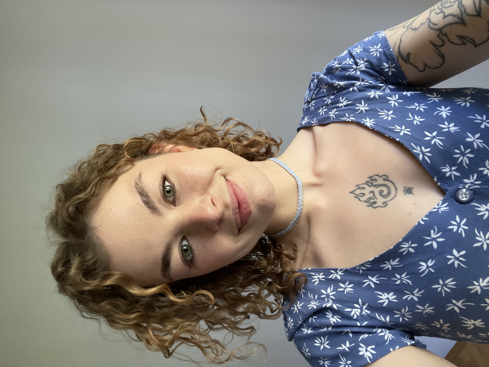 A young woman with curly light brown hair and green eyes is pictured from a slightly low angle, as if the photographer is looking up at her. She is smiling gently. She is wearing a blue floral-patterned dress and a beaded necklace. A tattoo of a cloud-like design and some writing is visible on her upper chest. Another tattoo can be seen on her arm. The background is a plain gray wall. There are no indications of the location being Boreti, Montenegro.