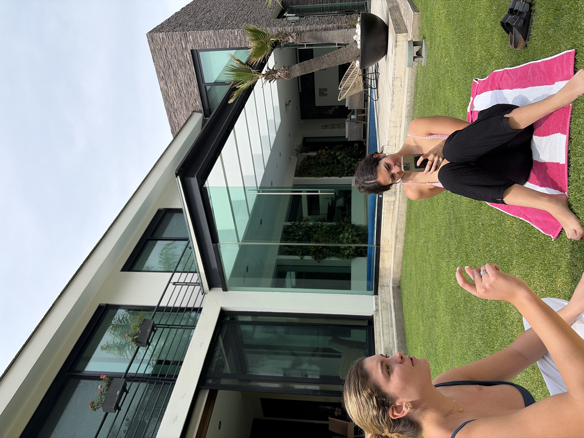 Here is a forensic description of the image:
The image presents two women in an outdoor setting. One woman is reclined on a pink and white striped towel, holding a mobile phone. The second woman is sitting beside her on the grass.
Behind them is a modern house with large windows, an overhanging roof, and a narrow swimming pool visible between the women. The area appears to be a lawn.
The location is Unknown, Unknown.