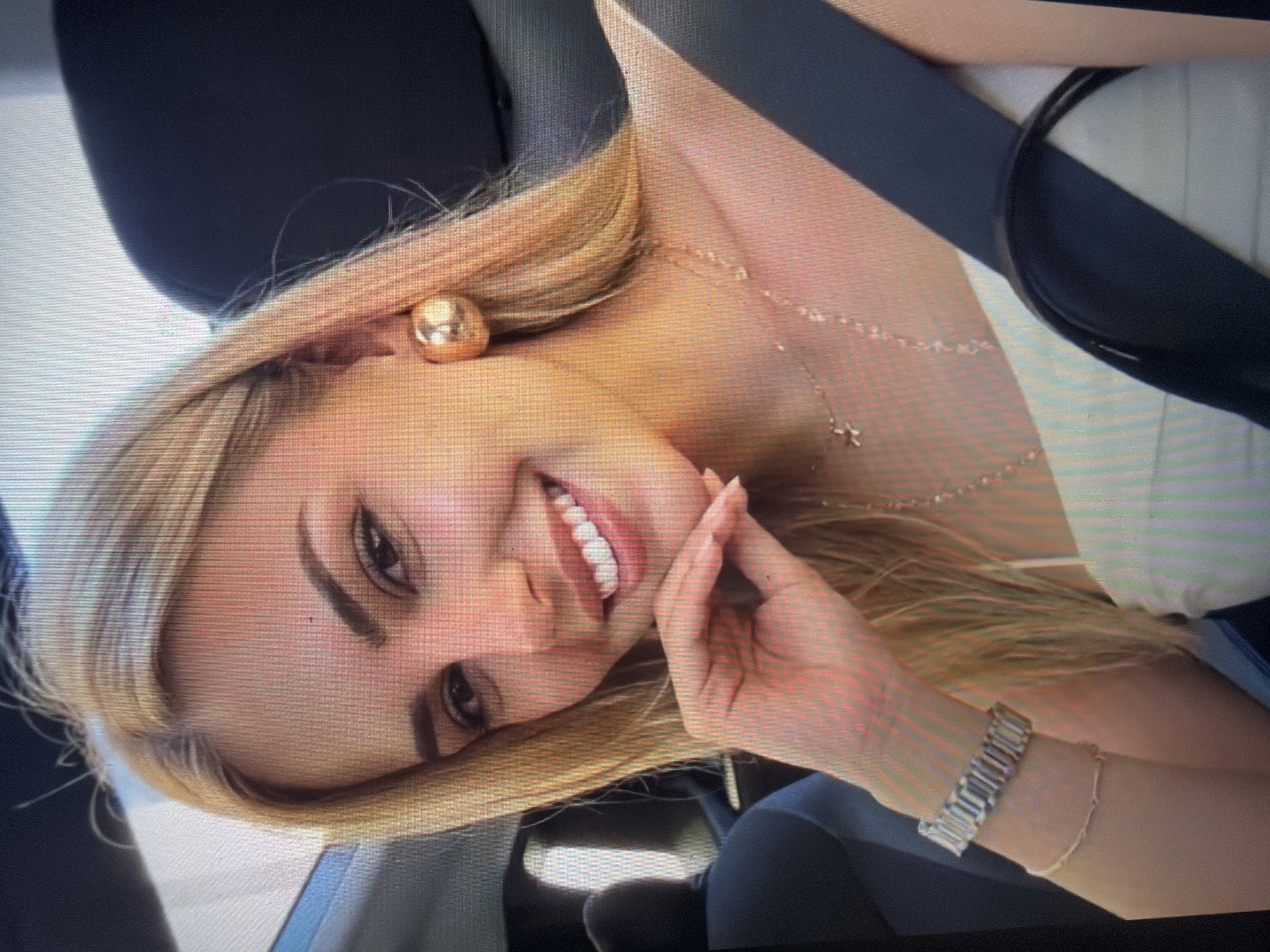 A close-up image, exhibiting significant pixelation and screen-capture artifacts, depicts a young adult female seated inside a vehicle. The scene is identified as the interior of a vehicle in Shumen, Bulgaria. The subject has long, straight blonde hair with visible darker roots at the part, and fine flyaway hairs near the forehead and ear. She is smiling broadly, revealing white, well-aligned teeth. Her left hand, with manicured nails, rests under her chin, her fingers gently curled. Her facial features include dark, well-defined eyebrows and visible eyeliner on her eyelids.
She is wearing a light-colored, possibly white, strapless or thin-strapped top. Adorning her right ear is a large, spherical, gold-toned stud earring. Around her neck, she wears multiple delicate gold-toned necklaces; one features a small, intricate pendant, possibly a cross. On her left wrist, she wears two bracelets: an upper wide, silver-toned bracelet composed of rectangular links, and a lower thin, light-colored chain bracelet. A dark-colored seatbelt is diagonally positioned across her chest and right shoulder. The vehicle interior is dark-toned, with a light-colored headliner and a partially visible sunroof above. The ambient lighting suggests a daytime setting.