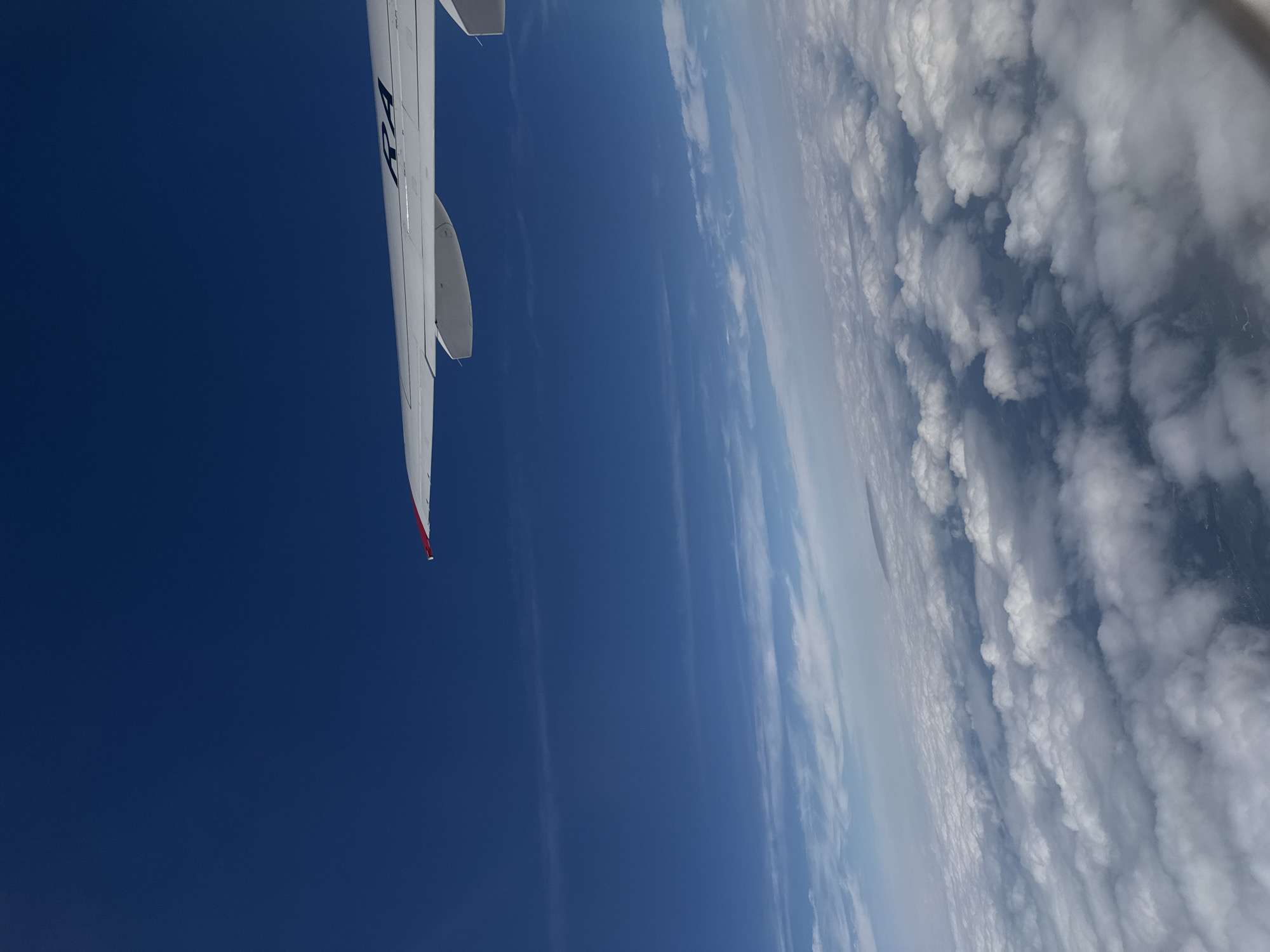 An aerial view shows a white airplane wing with the letters "RA" visible in dark blue lettering. The wing is positioned in the upper right portion of the image against a deep blue sky. Below the wing, a vast expanse of white clouds stretches across the horizon. The clouds appear fluffy and are layered, suggesting they are at a significant altitude. The perspective is from inside the aircraft, looking out. The geographical location of Staroe Mart'yanovo, Russia, is not discernable from the visual information provided in the image.