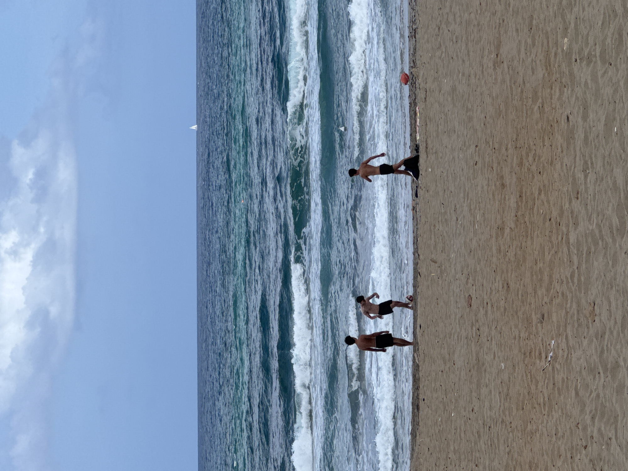Three young males are on a sandy beach, near the water's edge, on a cloudy day. Two of the males are actively engaged in a game of kicking a red ball. The third male stands facing the water. The sea is choppy with visible waves, and a distant sailboat can be seen on the horizon. The environment suggests a recreational beach scene, possibly in the city of Isola delle Femmine, Italy, given the description.