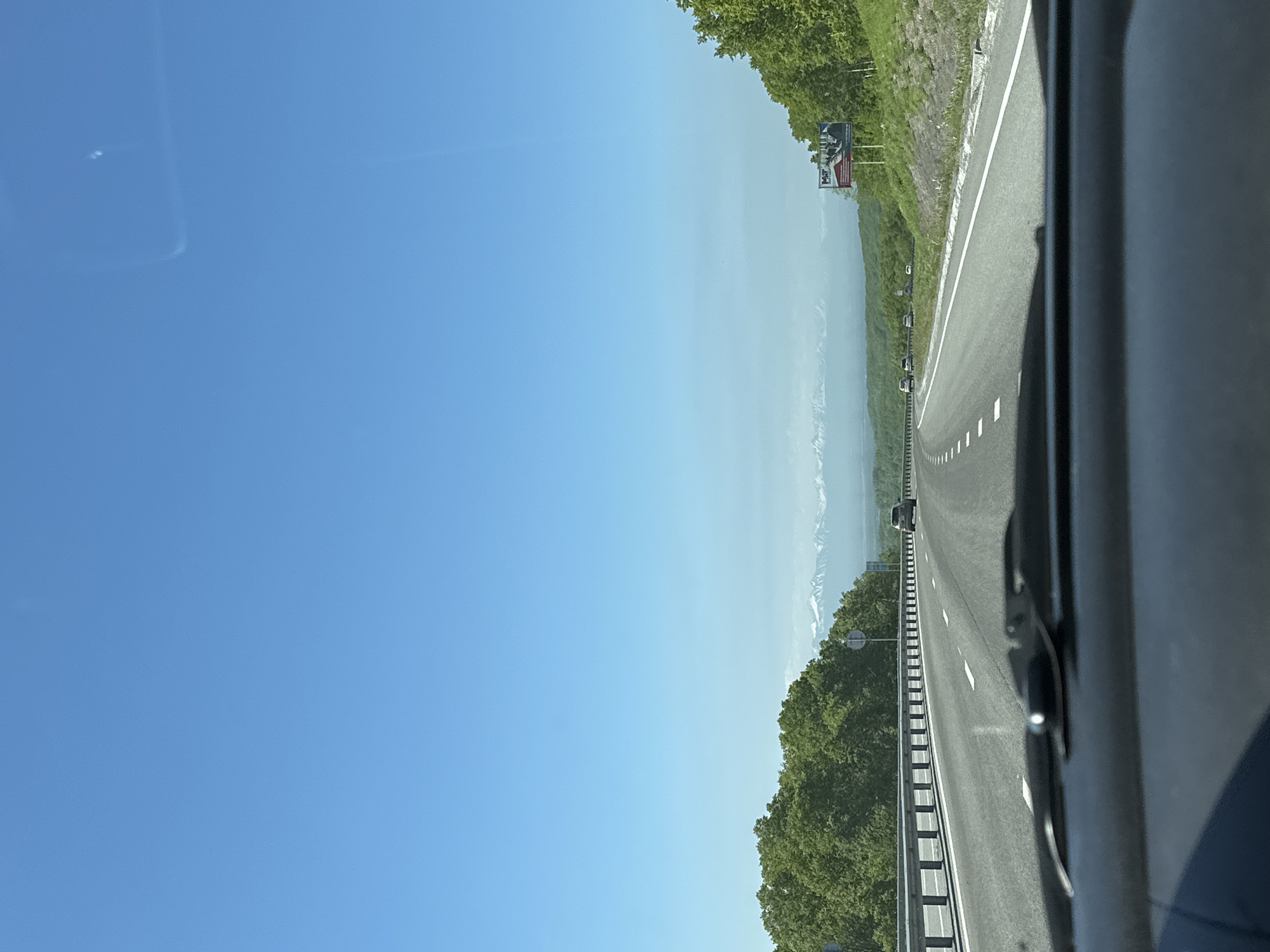 An eye-level, forward-facing shot captures a car's dashboard and windshield view of a highway on a clear, sunny day. The highway, with multiple lanes and white dashed lines, curves to the right, bordered by a metal guardrail and lush green trees on either side. In the distance, snow-capped mountains rise majestically under a vast, bright blue sky. Several cars are visible on the highway, moving in the same direction. A billboard with text and images stands on the right side of the road. The scene suggests a road trip through a scenic, mountainous region, possibly during spring or summer. The location is identified as the city of Svetly, Russia.