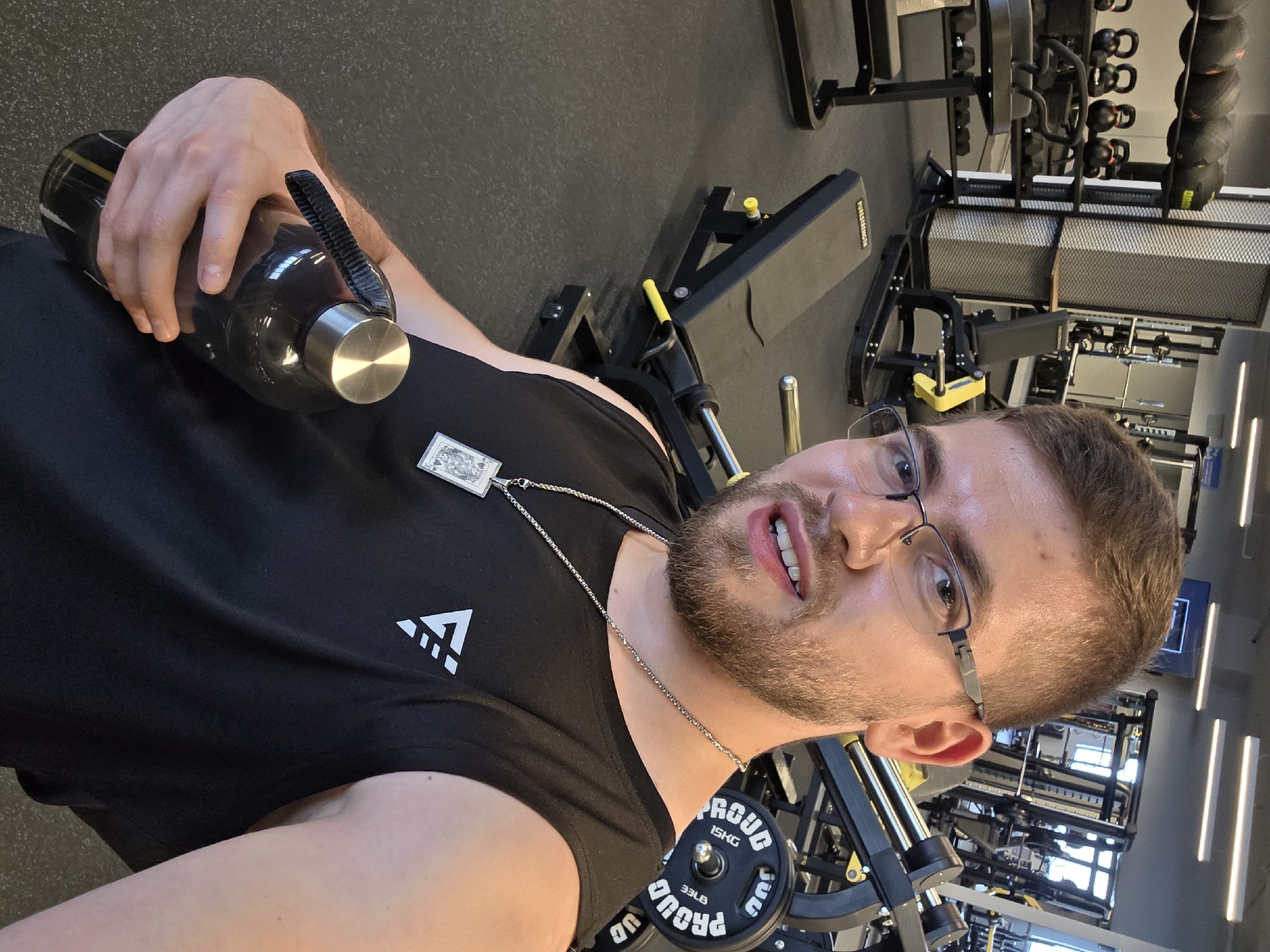 A fair-skinned man with short brown hair, a beard, and glasses is taking a selfie in what appears to be a gym. He is wearing a black tank top with a white logo on the left side and a silver chain with a pendant. He is holding a dark-colored water bottle in his left hand. The gym equipment is visible in the background, including weight machines and dumbbells. The weights read "PROUD 15KG 33LB". The location appears to be a gym in Plovdiv, Bulgaria.