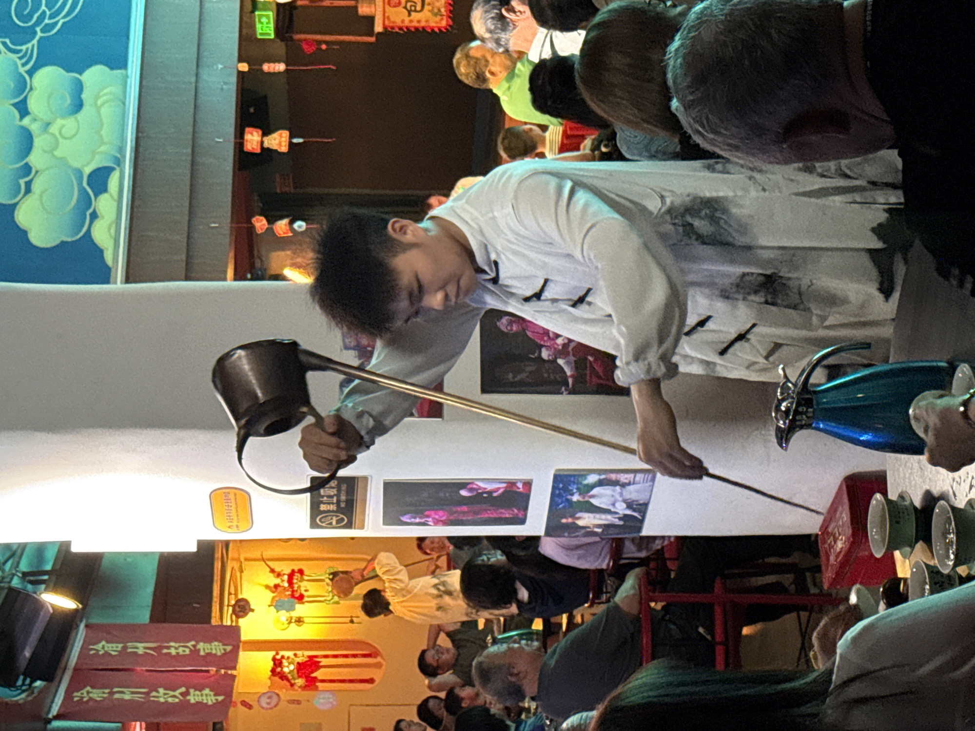 An entertainer is pouring tea at a tea house in the city of Yuzhong District, China. The entertainer is wearing a white robe with a black trim and pouring tea with a long, thin spout from a copper kettle into a cup. Several patrons are watching the performance. There are teacups, a table, a serving container, and decorations behind and above the performers and spectators.