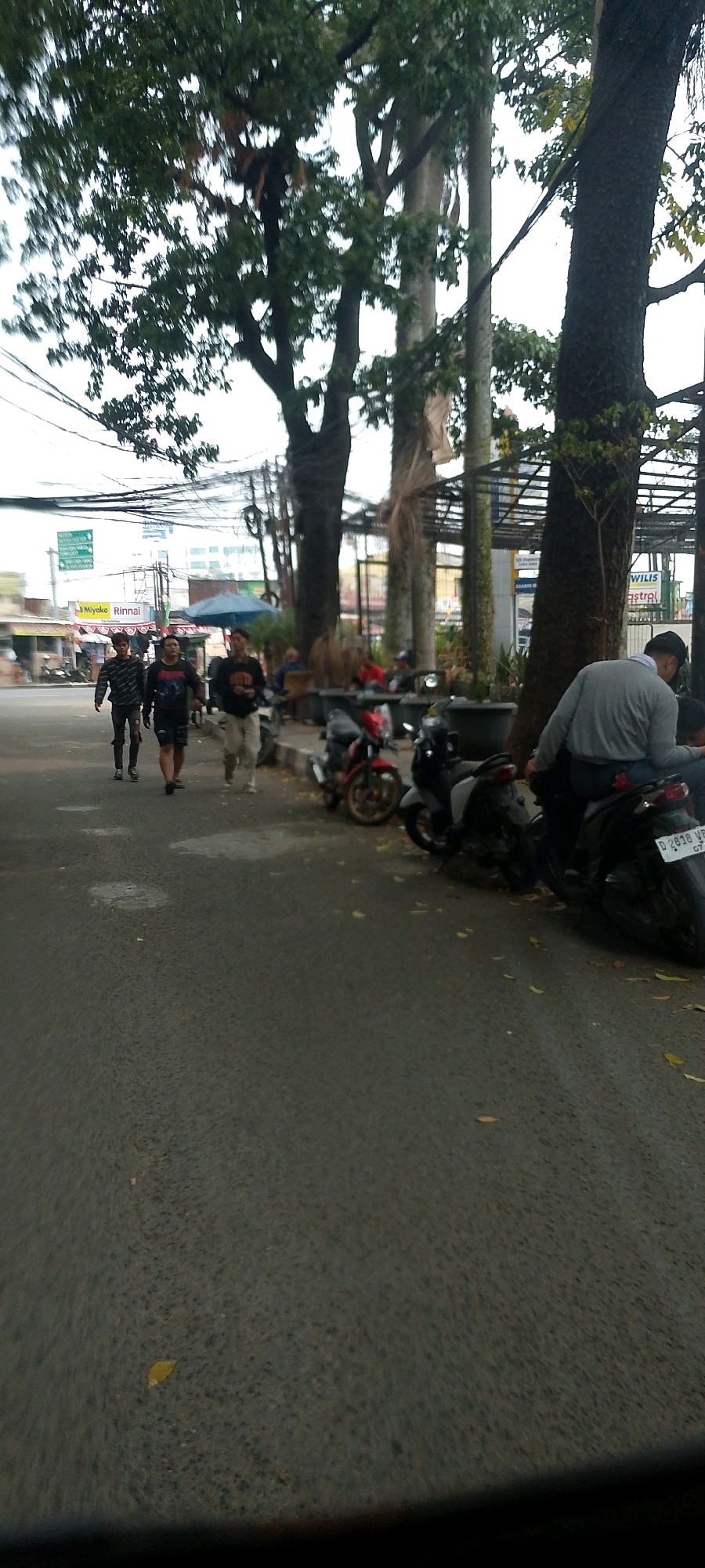The image shows a street scene in Bandung City, Indonesia. Three young men are walking on the right side of the road. The man in the foreground is wearing a black t-shirt and shorts, and the other two are wearing patterned shirts and pants. In the background, several motorcycles are parked along the sidewalk, and a person in a gray sweater is sitting on one of them. There are trees with dense foliage lining the street, and utility wires are visible overhead. Buildings and signs, including one for "Miyoko Rinnai", are present in the distance. The road surface appears wet in some areas, possibly indicating recent rain.