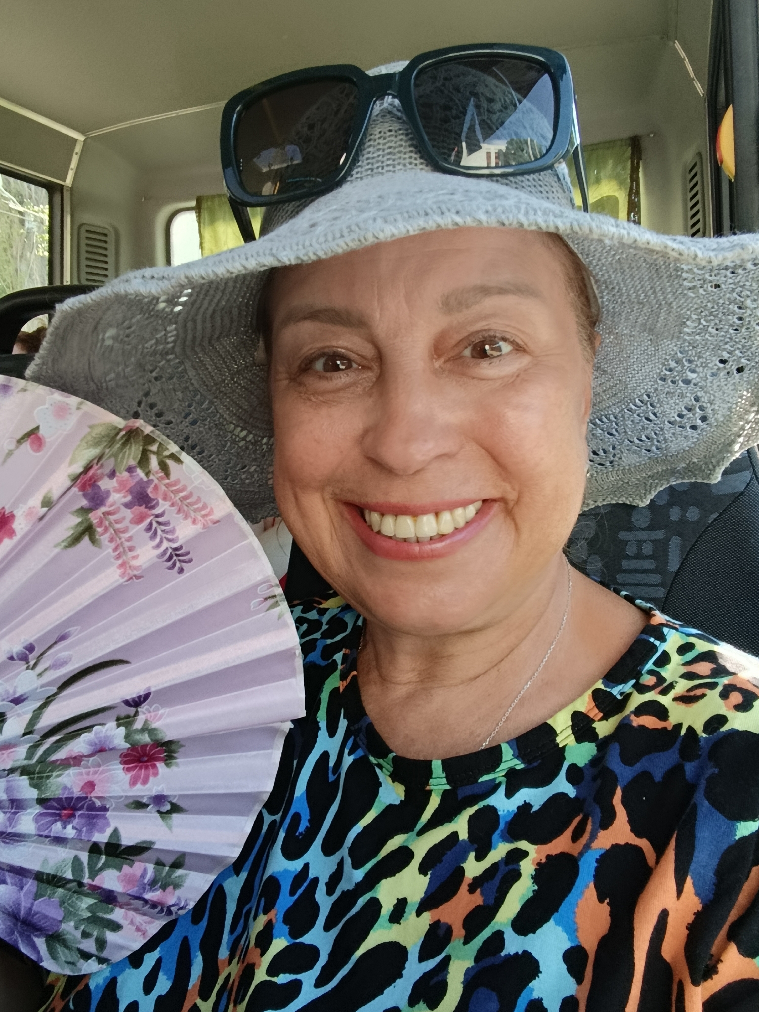A woman in her late 50s, with fair skin and brown eyes, is taking a selfie inside a vehicle. She is wearing a wide-brimmed, light grey hat and dark sunglasses perched on top of the hat. Her mouth is open in a wide smile, revealing her teeth. In her left hand, she holds a partially opened fan decorated with purple and pink floral patterns. She is wearing a brightly colored top with a leopard-print-like pattern in shades of blue, green, orange, and black. A thin silver necklace is visible around her neck. The background shows the interior of the vehicle, with a window on the left and green curtains on the right. The woman's right side is slightly obscured by the fan. The lighting suggests it is daytime, possibly with sunlight filtering in. There is no identifiable information about the location in Saratov, Russia.