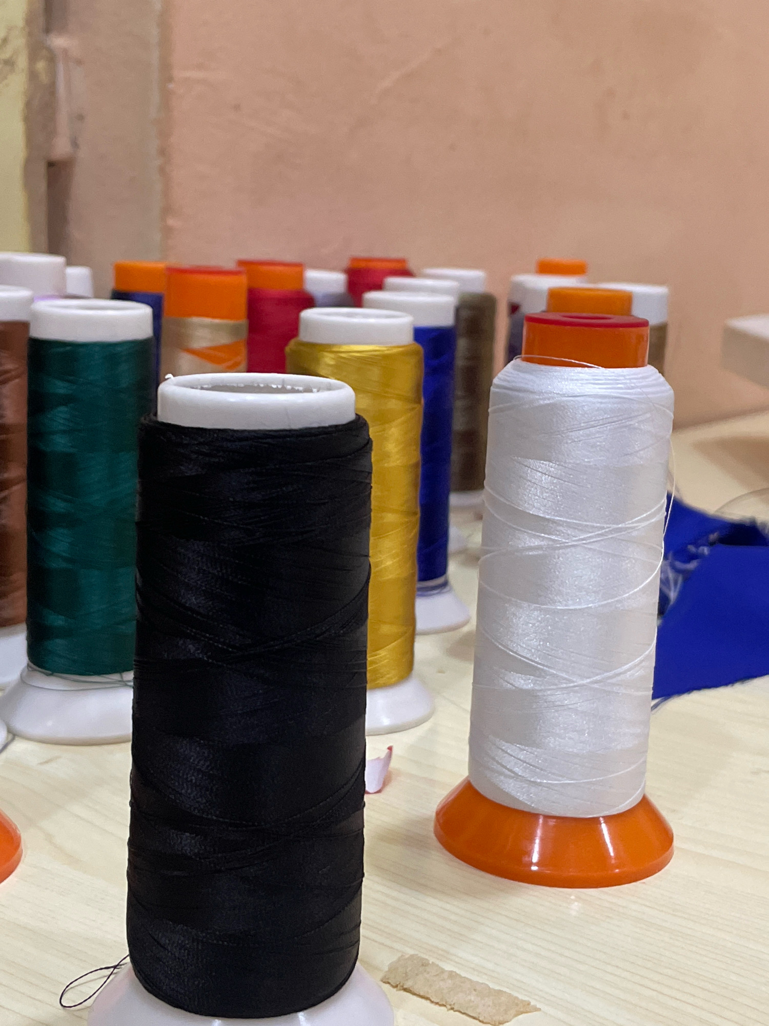 In Gura Top, Nigeria, a close-up shot focuses on a collection of spools of thread. The spools are arranged on a light-colored, possibly wooden surface. In the foreground, a large black spool of thread is prominently featured, standing on a white base. To its right, a white spool of thread, also on an orange base, is visible. In the background, several other spools of thread in various colors, including green, orange, red, yellow, and blue, are arranged in rows, some with white caps and others with colored plastic tops. The background wall is a muted, peachy-pink color. The lighting appears to be artificial and creates soft shadows. No people are present in the image.