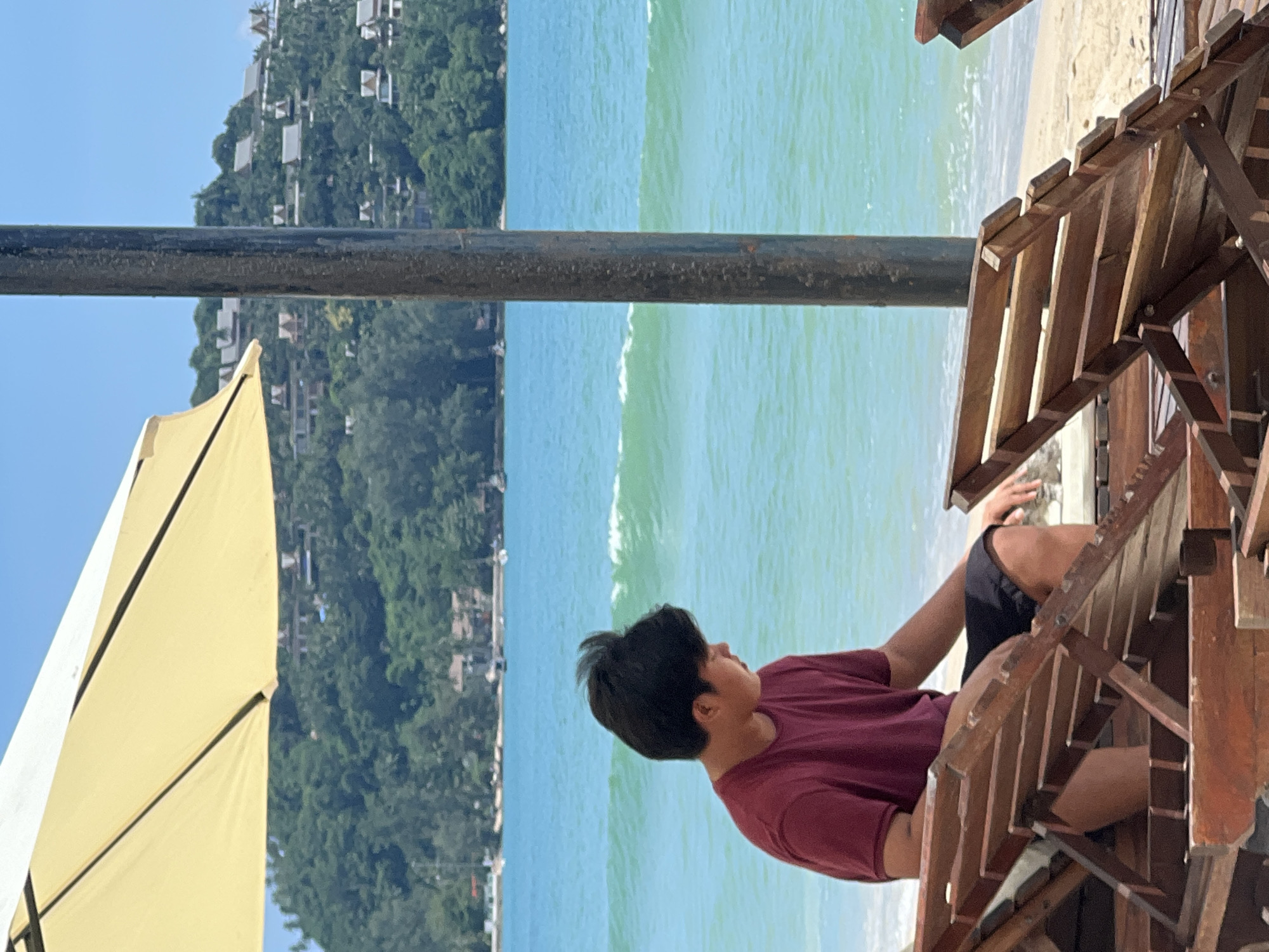 A young man in a maroon t-shirt and black shorts sits on a wooden beach chair, facing left, looking down. He is positioned under a large, partially visible yellow umbrella. Behind him, turquoise ocean waves gently break on a sandy beach. In the distance, a lush, green hillside is dotted with buildings. A dark, vertical pole stands to the right of the man, obscuring part of the background. The scene appears to be on a sunny day. The location is implied to be Choeng Thale, Thailand, known for its beaches.