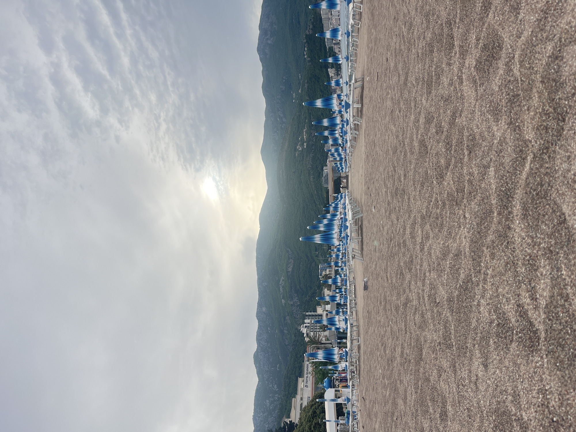 The image shows a beach in Boreti, Montenegro. The beach is sandy, and there are numerous blue and white striped beach umbrellas set up with lounge chairs. Behind the beach, a mountain rises, partly obscuring the sun. The sky is cloudy. Buildings are visible between the beach and the mountain.