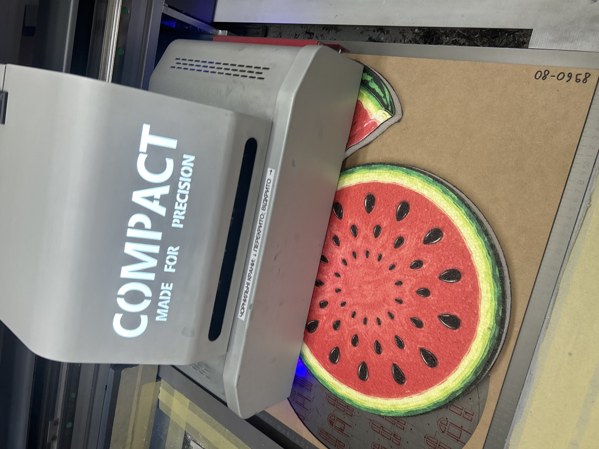 The image depicts a close-up view of a printing machine with the word "COMPACT" prominently displayed on its upper casing, followed by "MADE FOR PRECISION" below it. The machine is positioned over a light brown surface, likely a workbench or printing bed, on which two printed images of watermelon slices are placed. The larger watermelon slice is circular and detailed, with seeds and rind clearly visible. A smaller, partial watermelon slice is located to the right of the larger one. A blue light emanates from the printing head of the machine, casting a glow onto the watermelon print. To the left and below the watermelon prints, a portion of a ruler with measurements is visible. A label with Cyrillic text, "ЧОРНИЛЬНІ КРАНИ: 1 ПЕРЕКРИТО; ВІДКРИТО", is affixed to the side of the printing machine. The overall scene suggests an industrial or workshop environment, possibly related to printing or manufacturing. There is no information in the image to suggest a specific location within the city of Ternopil, Ukraine.