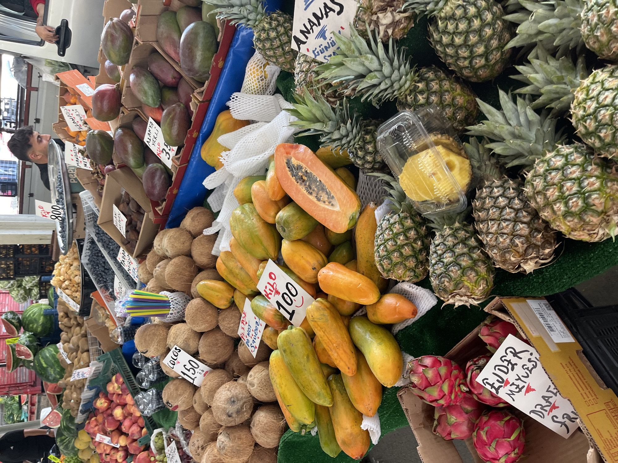 The image shows a fruit and vegetable stand in Alanya, Turkey. There is a variety of fruits, including mangos, dragon fruits, pineapples ("Ananas" in Turkish), coconuts ("Colos"), watermelons, papayas, and more. There are price signs written in Turkish. A man is visible in the background near the mangos. The fruits are arranged in piles and boxes. There are colorful straws near the coconuts. The sign "Alanya Yerli 35" indicates a product locally grown in Alanya.