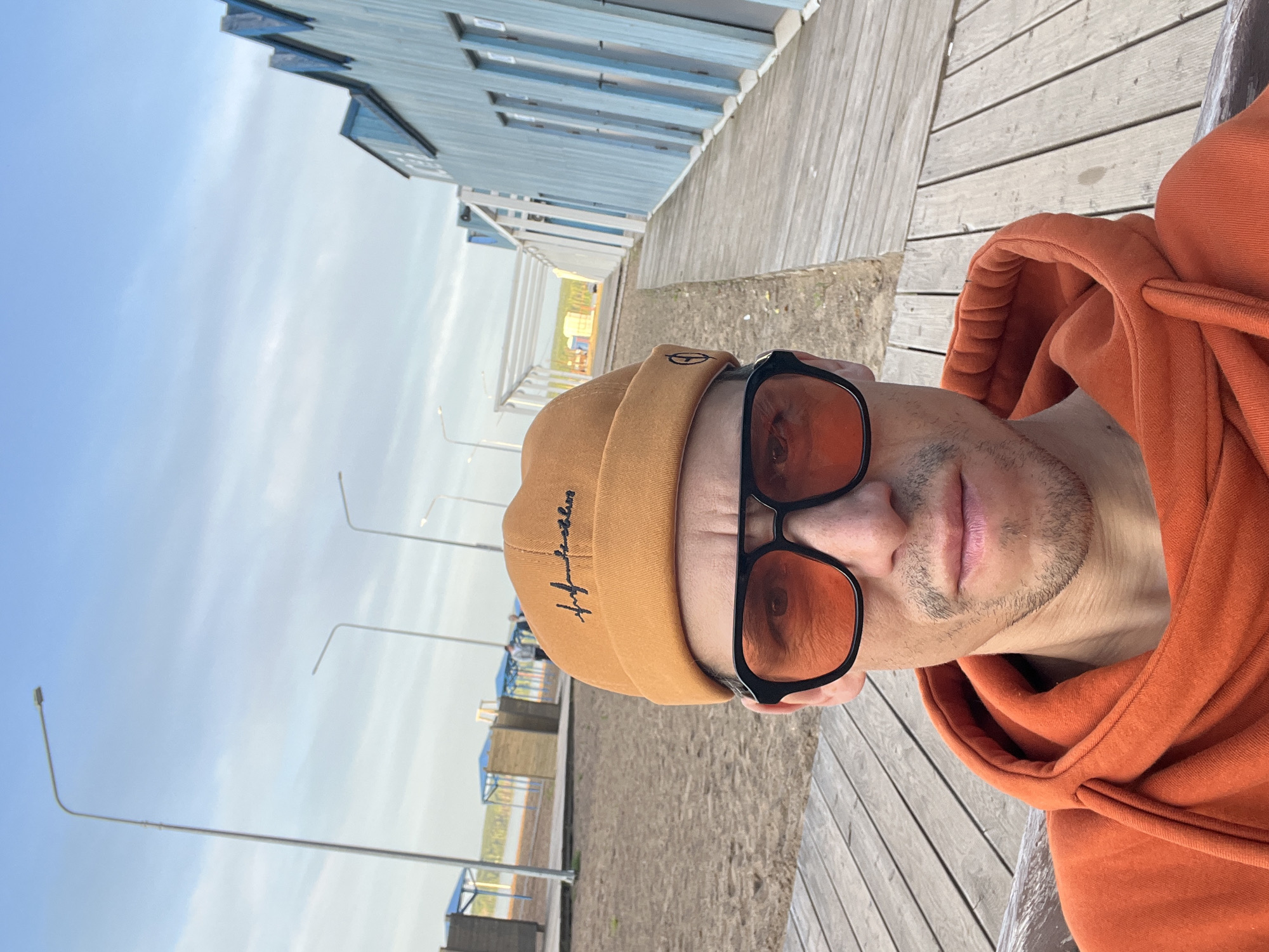 A man is captured in a close-up selfie, taken outdoors on what appears to be a wooden boardwalk or pier. He is seen from the chest up, with his head slightly tilted. He has light stubble on his chin and upper lip and is looking directly at the camera.
He wears a tan or light brown beanie, which features dark cursive embroidery on its front. He also wears black-framed sunglasses with distinct orange-tinted lenses. His attire includes an orange hooded sweatshirt, with the hood visible around his neck and shoulders.
The background shows an outdoor setting with a clear, light blue sky featuring faint clouds. To the man's left, a sandy or dirt ground extends towards several light blue structures, possibly beach huts or changing facilities, and beyond them, a distant landscape or body of water. To his right and behind him, more sections of the wooden boardwalk are visible, alongside a larger, light blue building with horizontal paneling and windows. Several streetlights with curved tops are sparsely placed against the sky.
The scene suggests daytime, possibly late morning or afternoon, given the clear sky and soft lighting. The overall environment appears to be a recreational area by the water.
Visible text: The beanie has embroidered text that reads "my future" in a dark, cursive font.
