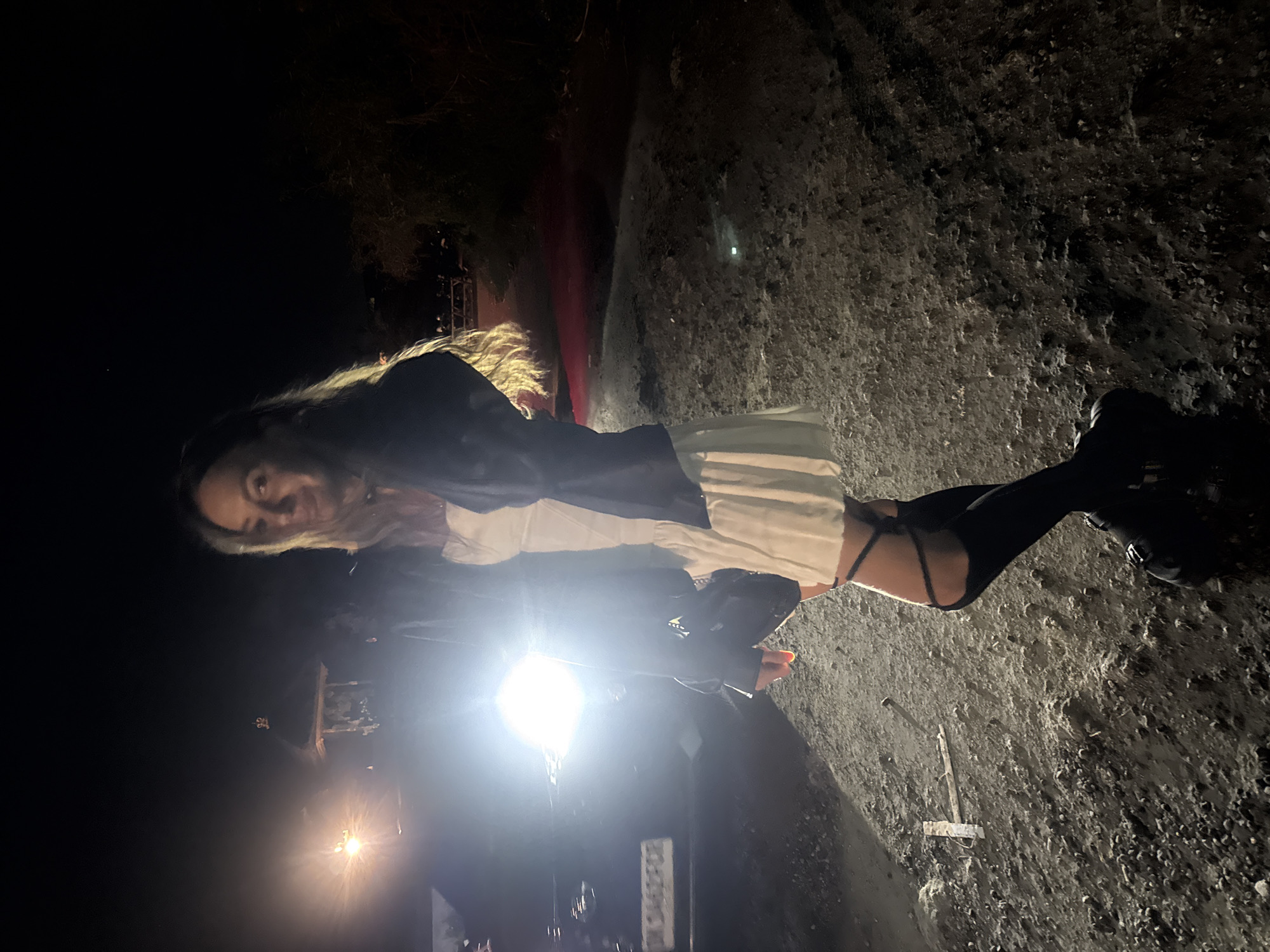 The photograph was taken at night in North Kuta, Indonesia. A woman stands illuminated by a bright light source, possibly a vehicle's headlights. She has long blonde hair and is wearing a dark jacket, a white top, and black thigh-high boots. She is positioned on a rough, asphalt-like surface with various debris scattered around. In the background, there are dark, indistinct shapes that appear to be vegetation and structures. The overall lighting is poor, with the light source creating strong contrasts and shadows.