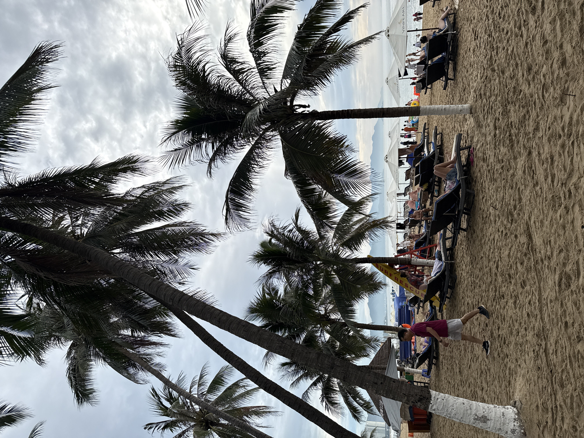 The image depicts a tropical beach scene in Nha Trang, Vietnam, likely at Vinpearl Land, as indicated by the umbrellas. The sky is overcast, with a light haze obscuring the distant mountains. The foreground is dominated by a sandy beach with numerous palm trees casting shadows. Several people are scattered across the beach, some lounging on beach chairs under umbrellas, while others are walking or standing. A man in a maroon shirt and grey shorts walks across the sand. Beach chairs and umbrellas are set up in rows. In the background, the ocean is visible, with a few people in the water. A yellow surfboard with "SURF RESCUE" written on it is propped up near a group of umbrellas. The overall atmosphere is relaxed, suggesting a vacation destination.