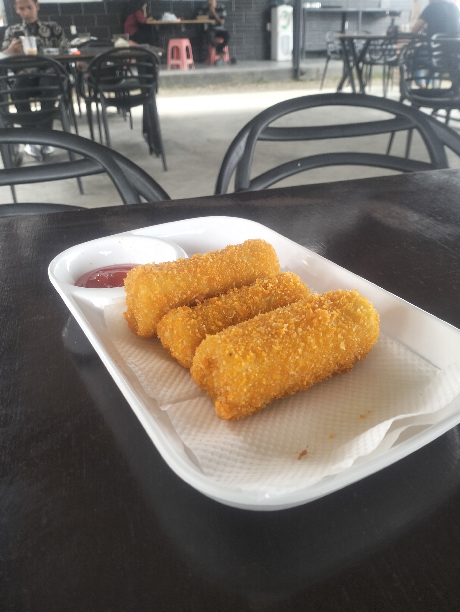A close-up shot shows a white rectangular plate filled with three golden-brown, breaded croquettes and a small portion of ketchup. The plate is placed on a dark, textured wooden table. In the background, the scene is blurred, showing a dimly lit, casual dining environment with people seated at tables and chairs. The establishment appears to be a cafe or restaurant. The image quality is low, making detailed identification of background elements challenging.