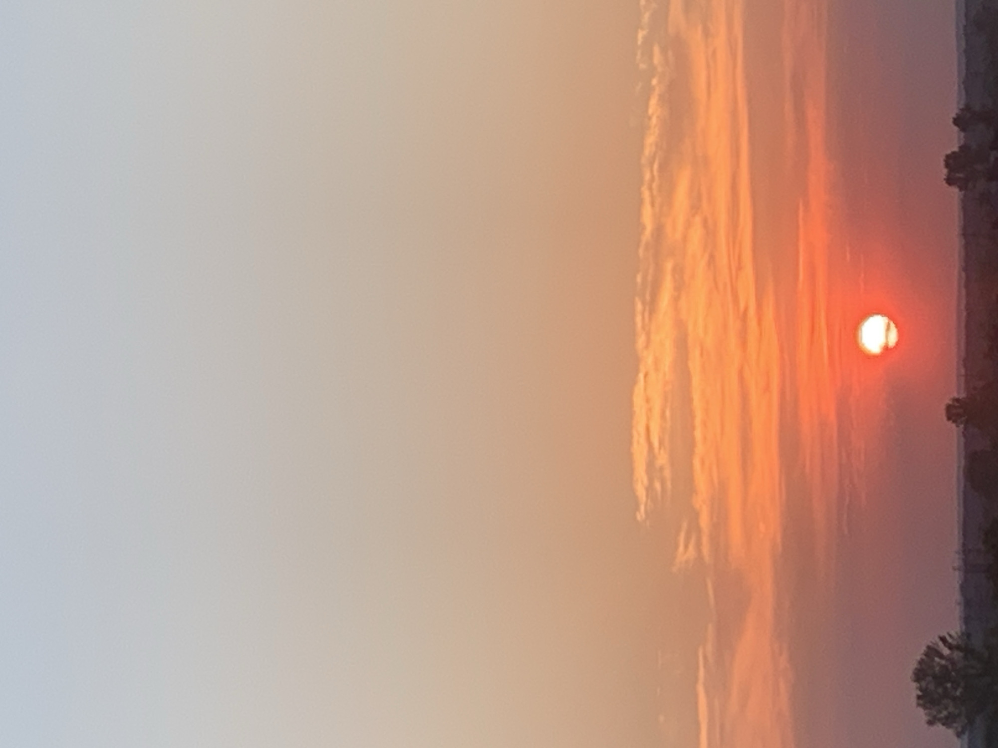 This image depicts a sunset over a flat, rural landscape, likely on the outskirts of Kolomyia, Ukraine. The sky displays a gradient of colors, transitioning from a pale blue at the top to a vibrant orange and red near the horizon, illuminated by the setting sun. Wisps of clouds are scattered across the sky, catching the warm hues of the sunset. The lower portion of the image shows a dark silhouette of trees and possibly distant structures against the colorful sky, suggesting a natural or undeveloped area. There are no people or discernible human activity visible in the scene. The overall impression is one of a serene, natural evening.