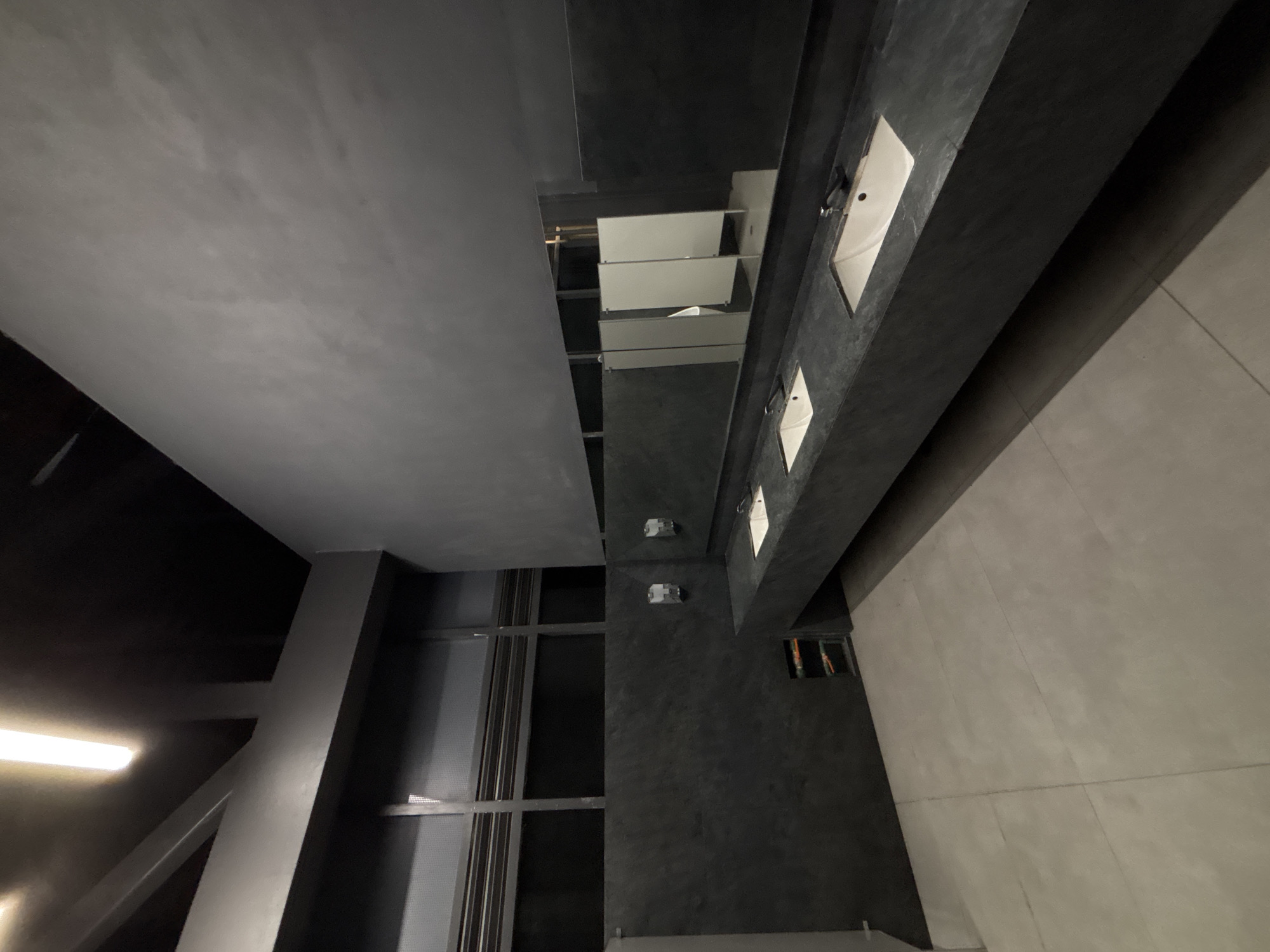The image shows an interior space with modern architectural design. A low-angle perspective includes a ceiling with a gray matte finish and a prominent structural support beam in the bottom left corner. The ceiling includes a modern lighting system, composed of linear lights in the dark area.
The right side of the image depicts a row of sinks embedded in a dark gray countertop with minimalist fixtures. Above the sinks are mirrors. The floor appears to be tiled in a similar gray tone.
The location is likely a public restroom or commercial space in Mexico City, Mexico, given the contemporary architectural style and common use of such materials in urban settings. No people or specific interactions are visible in the frame.