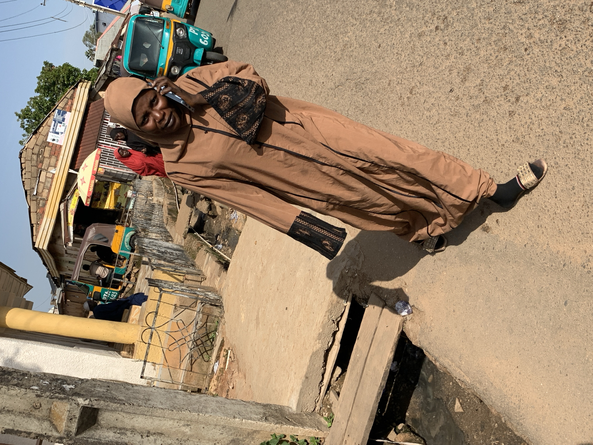 A full-body, slightly tilted shot captures a woman from the waist down, standing on a paved street. She is wearing a long, flowing brown abaya with black trim and lace detailing at the cuffs, a beige hijab, black socks, and light-colored slides. She is holding a black purse and a cell phone to her ear. Behind her, a turquoise and yellow three-wheeled taxi is parked on the street, and a small shop with a yellow awning is visible. The scene appears to be outdoors in a busy urban area, possibly a market or roadside with shops and traffic. The lighting suggests it is daytime, and the weather appears to be sunny. The ground is dusty, and there is a slight incline in the road. In the background, other vehicles and buildings suggest a bustling environment. The woman's expression is partially obscured by her phone, but she appears to be engaged in a conversation. The overall setting is a typical street scene in Gura Top, Nigeria, with elements of local commerce and transportation.