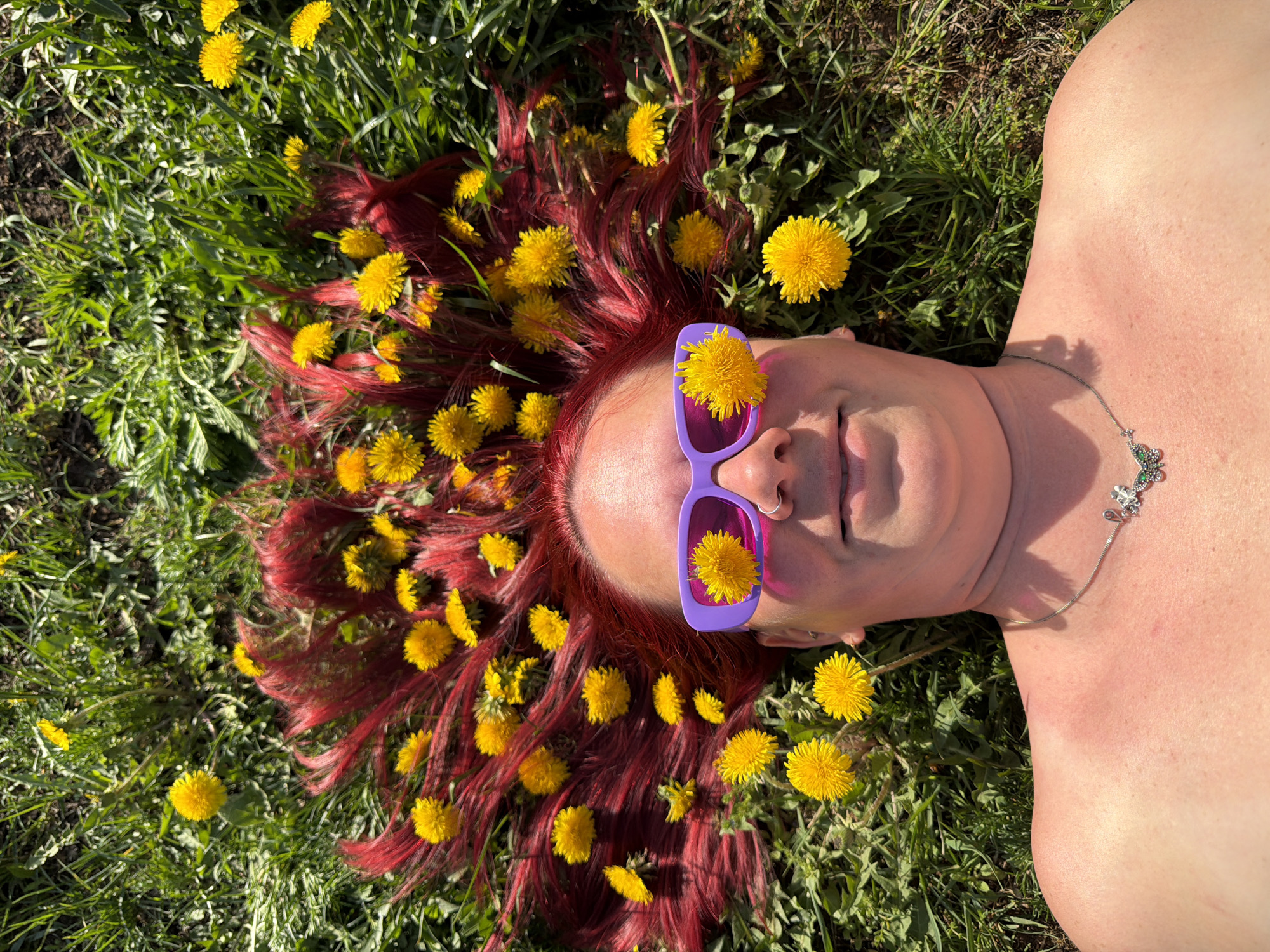 A person with red hair lies on their back in a field of green grass interspersed with yellow dandelions. The person is wearing purple sunglasses, with a dandelion stuck to each lens. The location of this image is unknown.