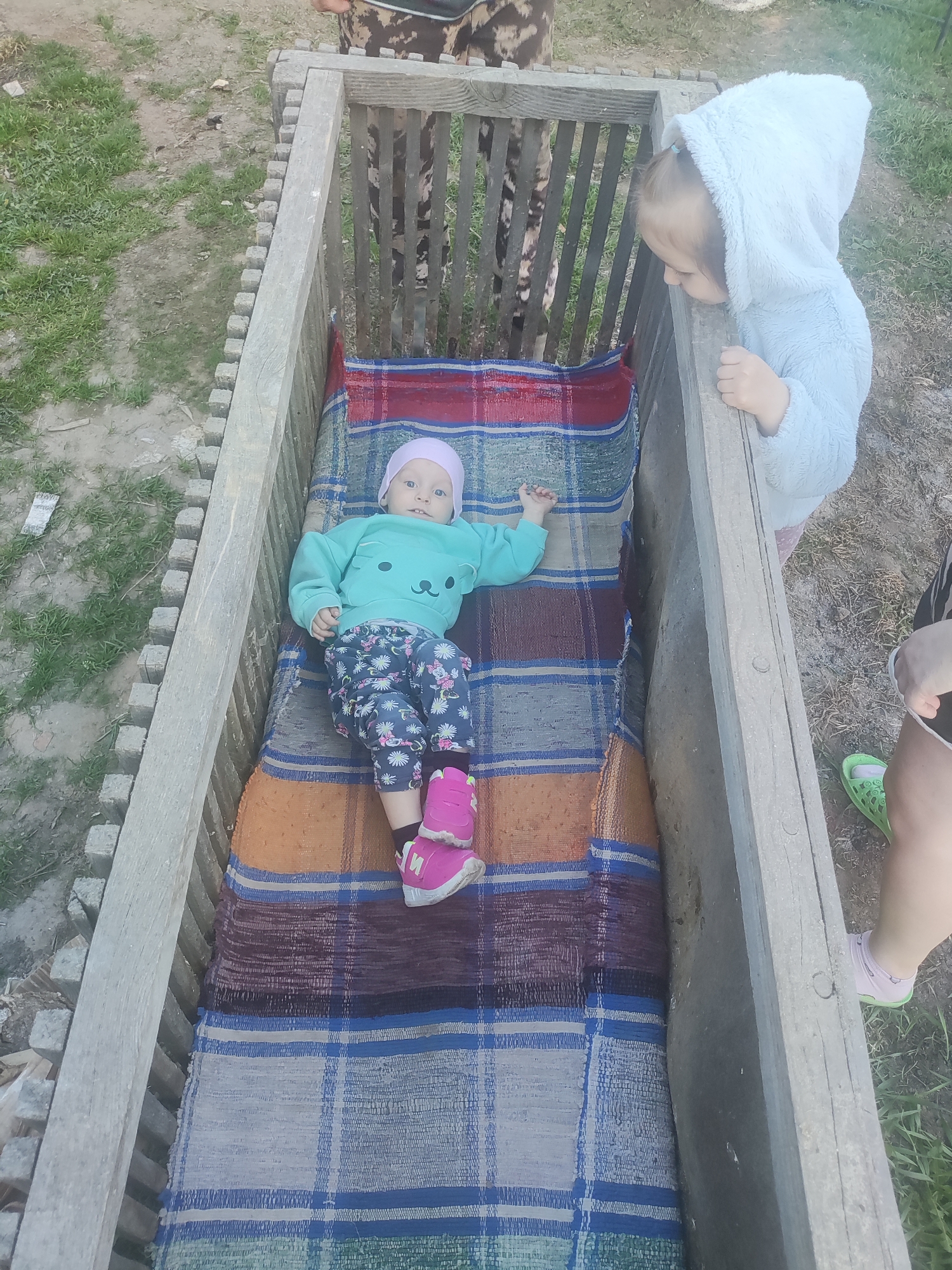 The image depicts a baby lying in a wooden trough-like structure, covered with a woven blanket. The baby is wearing a light blue sweater with a bear face on it, floral patterned pants, a pink hat, and pink shoes. Beside the trough stands a young child in a white hooded sweatshirt, looking down at the baby. Other people are partially visible around the trough. The scene appears to be outdoors, with grass visible in the background. The location is Nizhnyaya Gar', Russia.