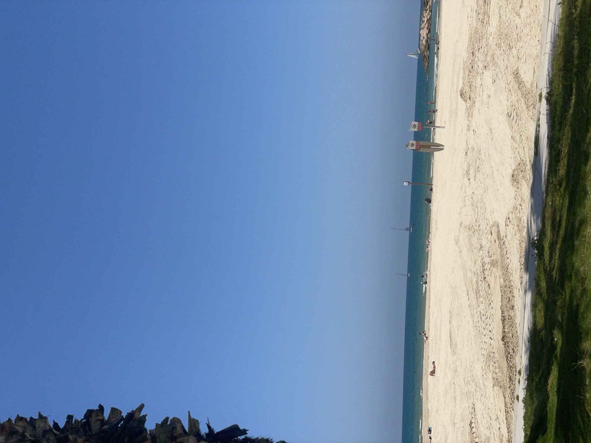The photograph depicts a beach scene. The beach is sandy with tire tracks and footprints, and there are signs along the shore. People are scattered along the beach, some sunbathing. Beyond the sand, there is water with a few sailboats in the distance. A grassy area borders the beach on one side, and the sky is a clear blue. Based on the location, this appears to be in Herzliya, Israel.