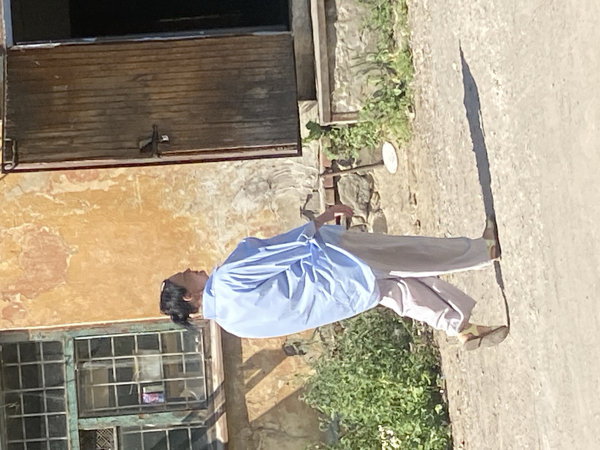 A person with dark hair pulled back in a bun walks away from the camera on a dirt path. They are wearing a loose, light blue collared shirt and light-colored pants. Their left foot is visible, wearing an open-toed sandal. The person is walking past a weathered building with an orange, textured wall. To the left of the building is a window with green-painted, gridded panes. To the right of the building is a large, wooden door that is partially open. Green foliage grows near the base of the building. The ground is composed of dirt and gravel. A white, circular object is visible on the ground to the left of the person's feet. A long shadow is cast on the ground to the right of the person, indicating a bright light source.