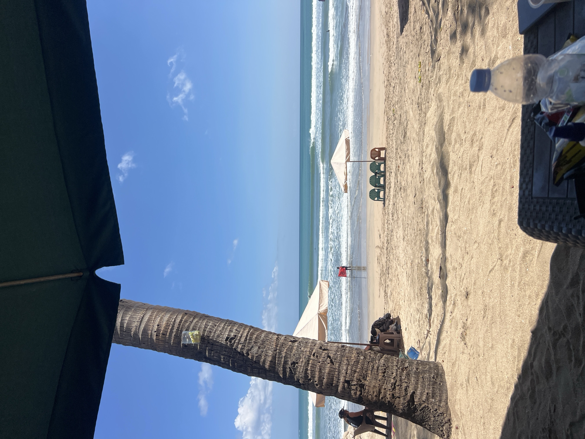 A beach scene in Kuta, Indonesia is captured from a shaded area. The foreground is dominated by a large palm tree on the left and the edge of a dark green umbrella and a wicker table with a plastic water bottle, suggesting a seating area. The sand on the beach is light brown and textured with shadows cast from overhead.
Further back, several beach chairs are visible under a beige umbrella. A person in a red shirt stands in the shallow water, facing away from the camera, near a red flag. The ocean is a vibrant turquoise with moderate waves and a clear blue sky with scattered white clouds above. Several surfers can be seen in the water. The overall atmosphere is a sunny day at a tropical beach.