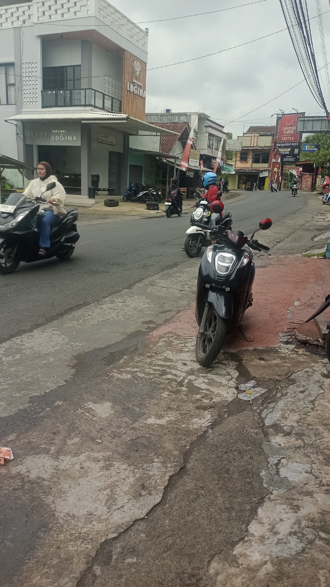 The image depicts a street scene in Karyamandala, Indonesia, under an overcast sky. A black scooter is parked on a red-stained concrete area in the foreground. To the left, a woman wearing a beige jacket and blue jeans rides a black scooter on a paved road. Behind her, several other scooters are parked or in motion.
Further down the street, a white scooter with a blue helmet rider is visible, as is another scooter with a red helmet rider. Buildings line the street, including a multi-story structure with a sign displaying "TOKO MAS LUGINA" (Lugina Gold Shop) and another building with a "DJARUM SUPER KRETEK" advertisement. The environment appears to be a commercial area with various shops and businesses. The road surface is asphalt, with patches of concrete and some debris scattered on the ground. Electrical wires are visible strung across the top of the frame.