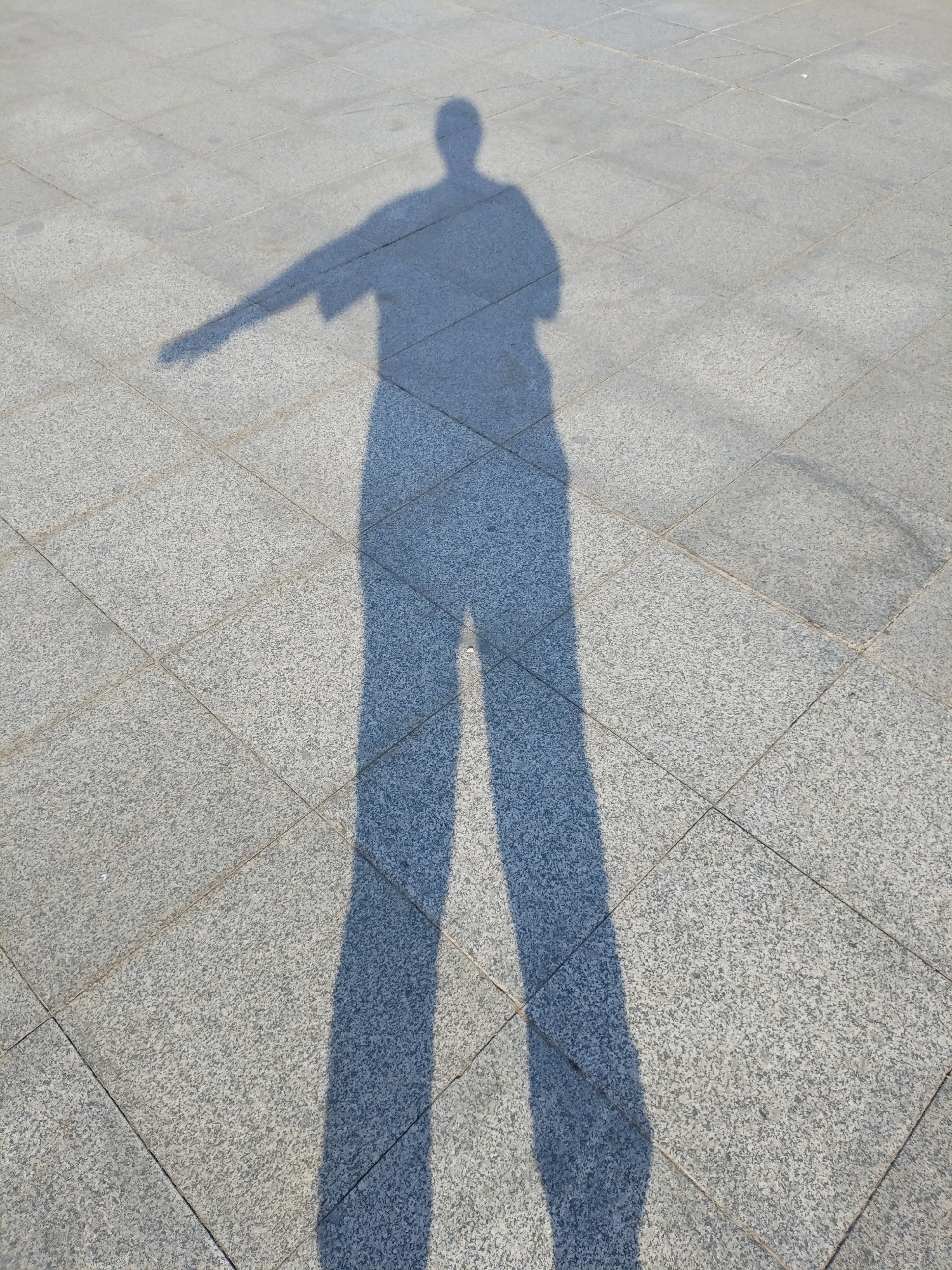 A shadow of a person with arms outstretched is cast on a granite-tiled pavement. The person is standing and appears to be in motion, possibly dancing or striking a pose. The shadow's left arm is extended horizontally, and the right arm is slightly bent at the elbow. The legs are spread apart, creating a dynamic silhouette. The pavement is composed of square, light-colored granite tiles with visible dark speckles. The sunlight is strong, casting a sharp, well-defined shadow. The scene suggests an outdoor location in Komsomolsk-on-Amur, Russia, during daylight hours.