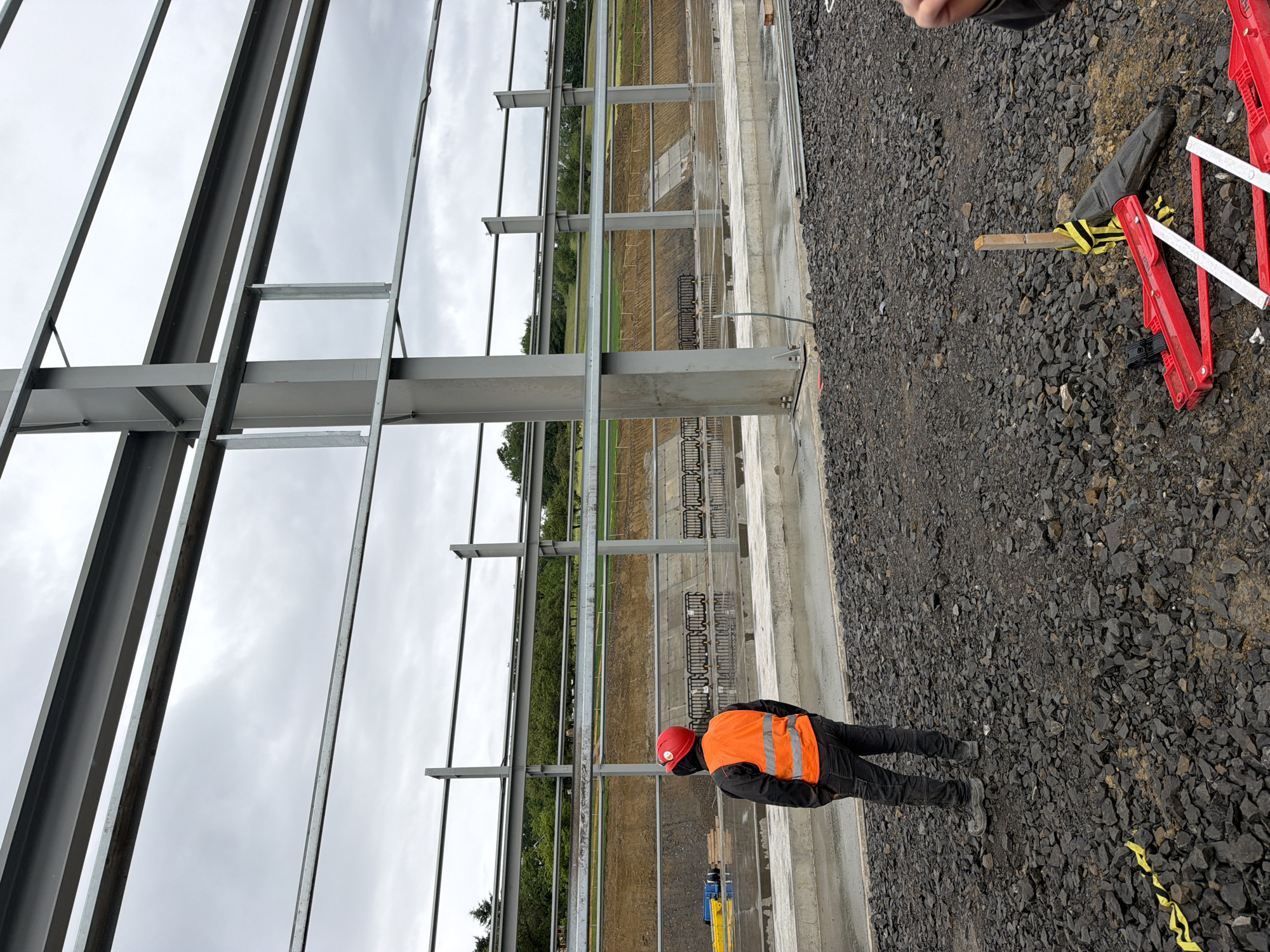 In Stróża, Poland, a construction worker stands on a gravel surface, facing away from the camera. The worker is wearing a red hard hat and a bright orange safety vest over dark clothing and work boots. Behind the worker, a large, unfinished steel structure is visible, with horizontal beams extending across the frame. The background shows a graded slope with dark soil and some vegetation, indicating ongoing construction work. The sky is overcast. In the foreground, near the worker's feet, a yellow caution tape lies on the gravel, and a pair of red construction jacks and a black object are also present.