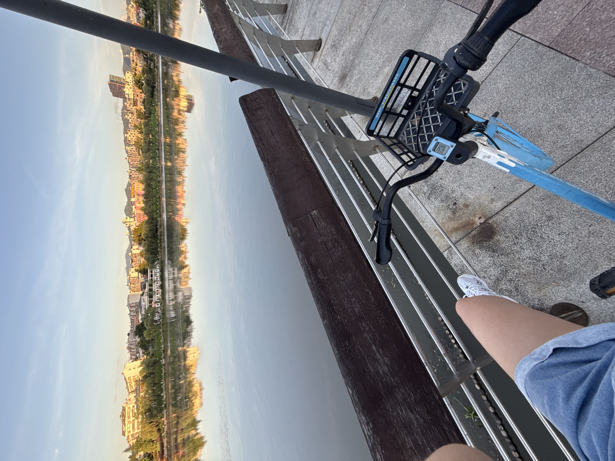 The image depicts a bright, outdoor scene featuring a person's leg and a bicycle next to a body of water, likely a lake or river, with a city skyline in the background. The time of day appears to be late afternoon or early evening, indicated by the warm lighting and long shadows.
The foreground is dominated by the front of a blue and white shared bicycle, equipped with a black basket and a digital display or QR code reader. A person's right leg, clad in denim shorts and a white sneaker, is visible on the left side of the image, positioned as if they are about to ride the bicycle. The ground is paved with stone tiles.
The middle ground shows a wooden and metal railing along the edge of the water. The water itself is calm, reflecting the sky and the cityscape.
The background presents a city skyline with numerous buildings of varying heights and architectural styles. Trees and greenery line the waterfront. The sky is a clear blue with some wispy clouds. The overall atmosphere is serene and suggests a recreational setting within a city. The presence of a shared bicycle implies an urban environment like Yiwu, China, known for its extensive public transportation options and parks.