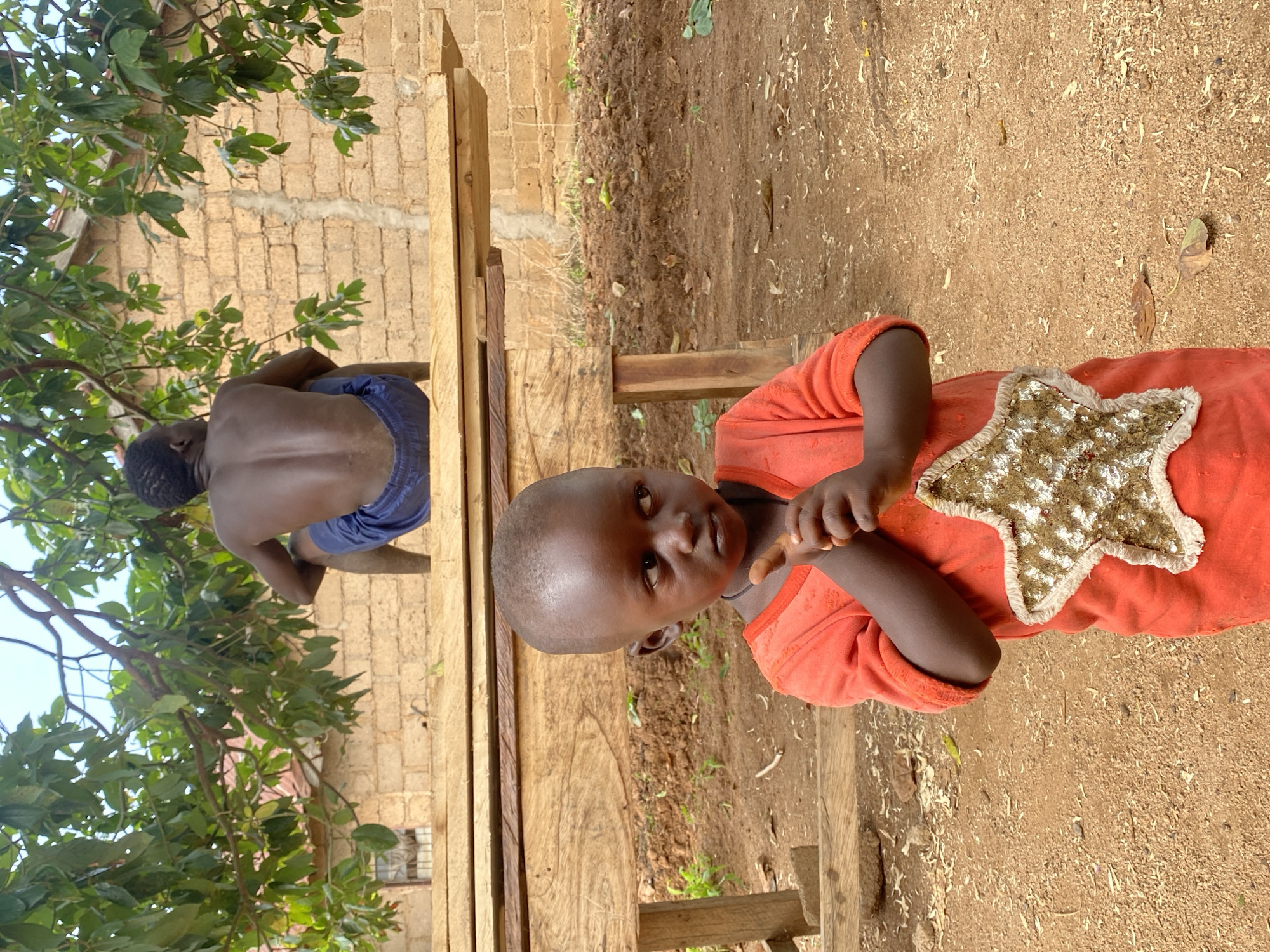 In Riyom, Nigeria, a young child wearing an orange dress with a star-shaped sequined appliqué stands outdoors on a dirt ground. The child is looking at the camera with their fingers intertwined. Behind the child, there is a wooden bench or structure composed of planks, and a man in blue shorts is squatting next to the structure with his back facing the camera. A tree with green foliage is to the left of the frame. The backdrop includes a brick building.