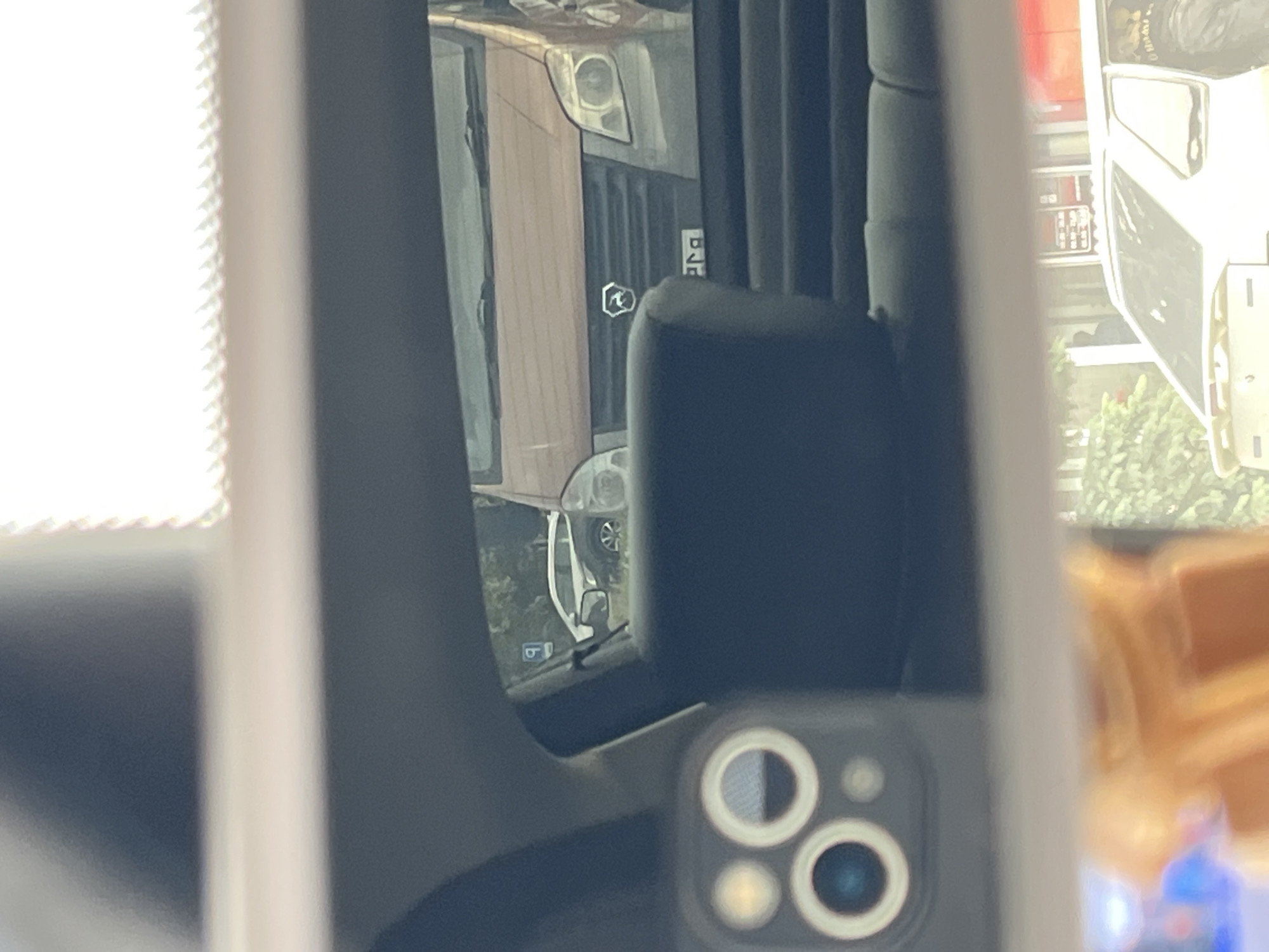 The image is a reflection in a car's rearview mirror, showing several vehicles and a building. In the foreground, a smartphone with multiple lenses is visible on the left. In the middle ground, a dark blue car seat is prominent, and behind it, a light brown van with a grille and a badge is parked. To the left of the van, a white SUV with a parking sign above it is visible. In the background, a white car is parked in front of a building with windows and signs. The scene appears to be in an urban environment with some greenery visible. The license plate on the van appears to be in Cyrillic script.