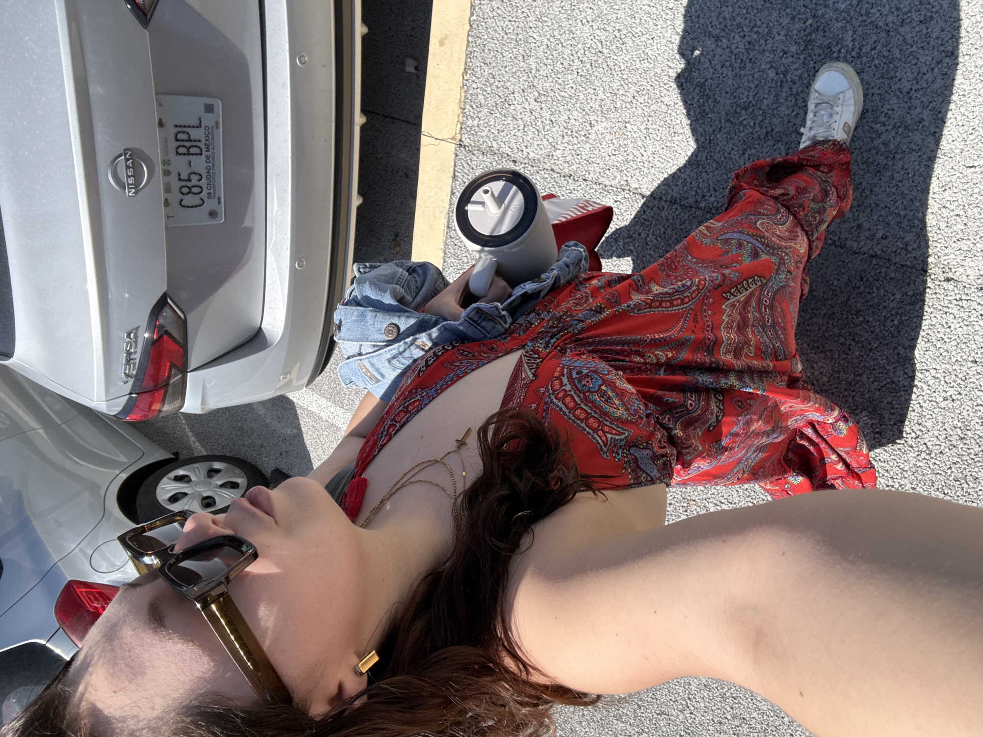 This image captures a person relaxing outdoors in what appears to be a sunny parking lot or paved area in Mexico City. The shot is taken from a low angle, looking up at the subject, who is lying down or leaning back.
The main subject is a person, likely female, dressed in a vibrant red, paisley-patterned dress or jumpsuit. A light blue denim jacket is casually placed or worn around her mid-section. She wears white sneakers with a dark sole, and a brand logo is subtly visible on the side of one shoe. Accessories include large, brown-framed sunglasses, two delicate gold chain necklaces (one with a small cross pendant), and rectangular gold earrings. Her long, dark hair is visible around her face and shoulder.
Beside her, a white tumbler with a black rim and a straw is visible, held or resting near her arm. In the background, two silver cars are parked. The car closest to the subject is a Nissan Versa, clearly indicated by the "NISSAN" logo and "VERSA" text on its rear. This car bears a Mexican license plate displaying "C85-BPL" and "09 CIUDAD DE MÉXICO" (09 Mexico City), along with the text "TRANSPORTE PRIVADO NOTORIO". The rear section of another silver car is partially visible to the left.
The bright sunlight casts strong shadows, suggesting it is midday or early afternoon on a clear, sunny day. The ground is a light-colored asphalt or similar paved surface. The overall impression is one of leisure and relaxation in an urban outdoor setting.