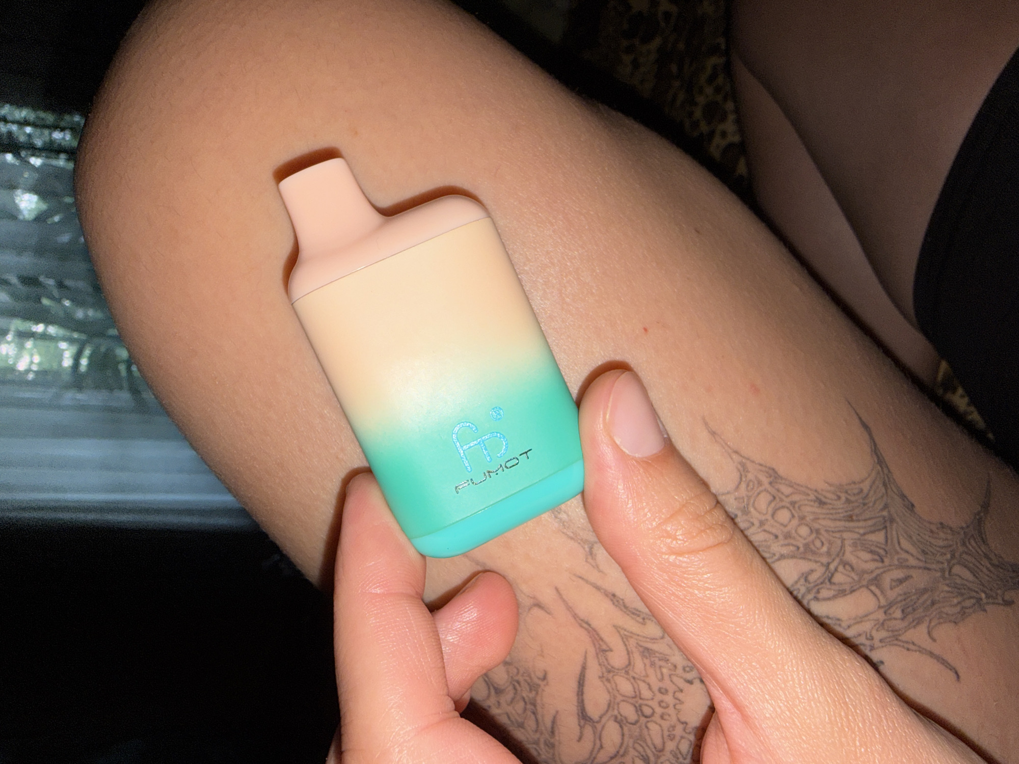 A person's hand holds a light-colored vaping device with a gradient of green to pink. The device has a logo that appears to be "FB" in blue. The person's skin is visible, and there is a tattoo on their forearm. The background is dark with some light filtering through what appears to be curtains. The overall scene is dimly lit.