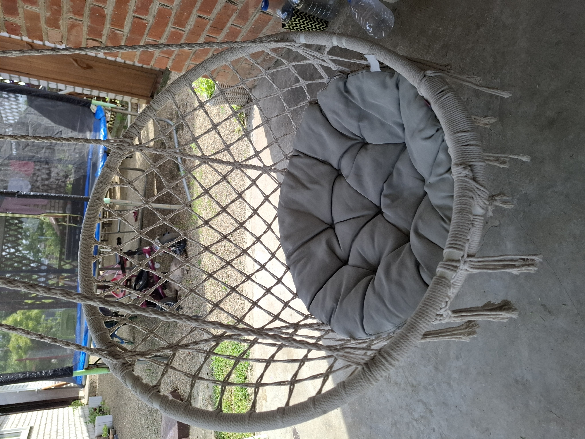 A close-up, eye-level shot shows a hanging chair with a gray cushion, suspended by thick ropes. The chair is woven with a diamond pattern, and fringe hangs from its bottom. In the background, a trampoline is visible, as are two plastic bottles and a brick wall. The scene is set on a concrete patio with gravel and grass. The city of Korenovsk, Russia, is implied by the context. No people are clearly visible, though a small, blurry figure is discernible behind the chair. A cat is lying on the ground in the background.