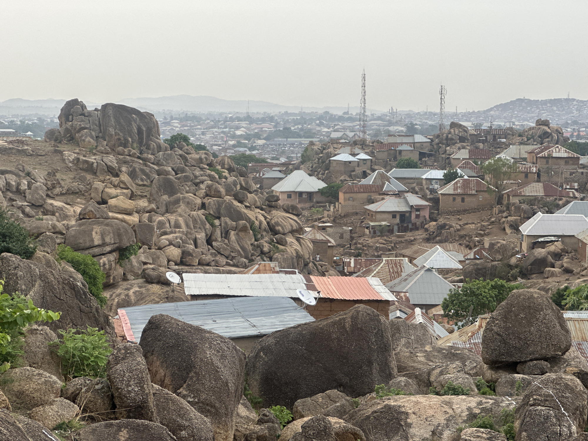 The image shows an elevated view of a section of Jos, Nigeria. The foreground is dominated by large, weathered granite boulders. Scattered among and beyond the rocks are houses with various metal roofs, some rusted, others painted. Satellite dishes are visible on some roofs. In the midground, more houses extend into the distance, blending into the sprawling city. Communication towers rise above the urban landscape in the distance. A line of hills is barely visible on the horizon under an overcast sky. There are no visible people. The environment appears rocky and arid.