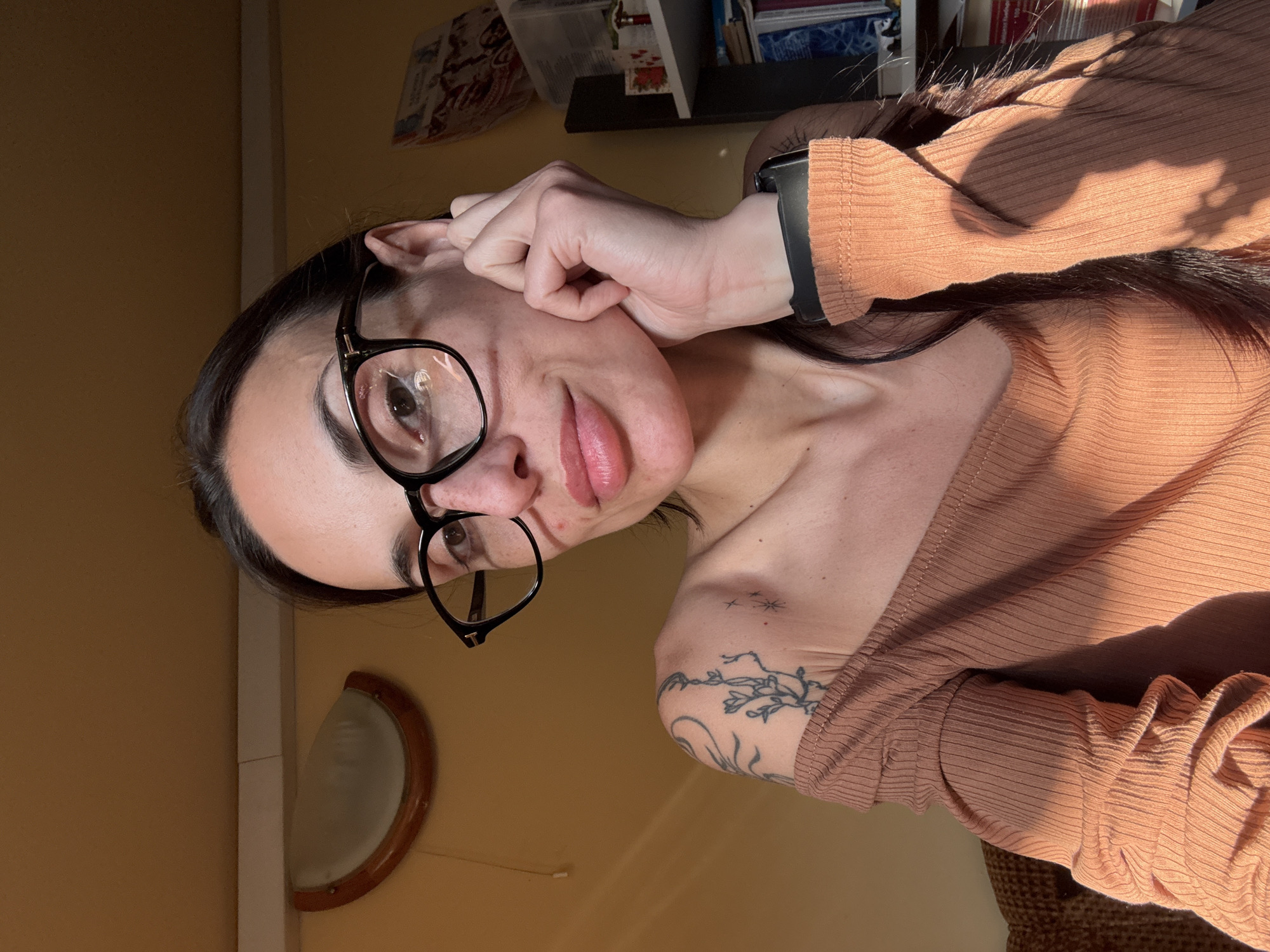 A woman is photographed in a brightly lit indoor setting. She wears black-rimmed glasses and a brown, off-the-shoulder, ribbed shirt. Her dark hair is pulled back. She has a tattoo on her right shoulder. A black smartwatch is on her left wrist. The background includes a yellow wall, a semi-circular light fixture, a shelf with books, and what appears to be wicker furniture. There is no evidence to support that this picture was taken in Pleven, Bulgaria.