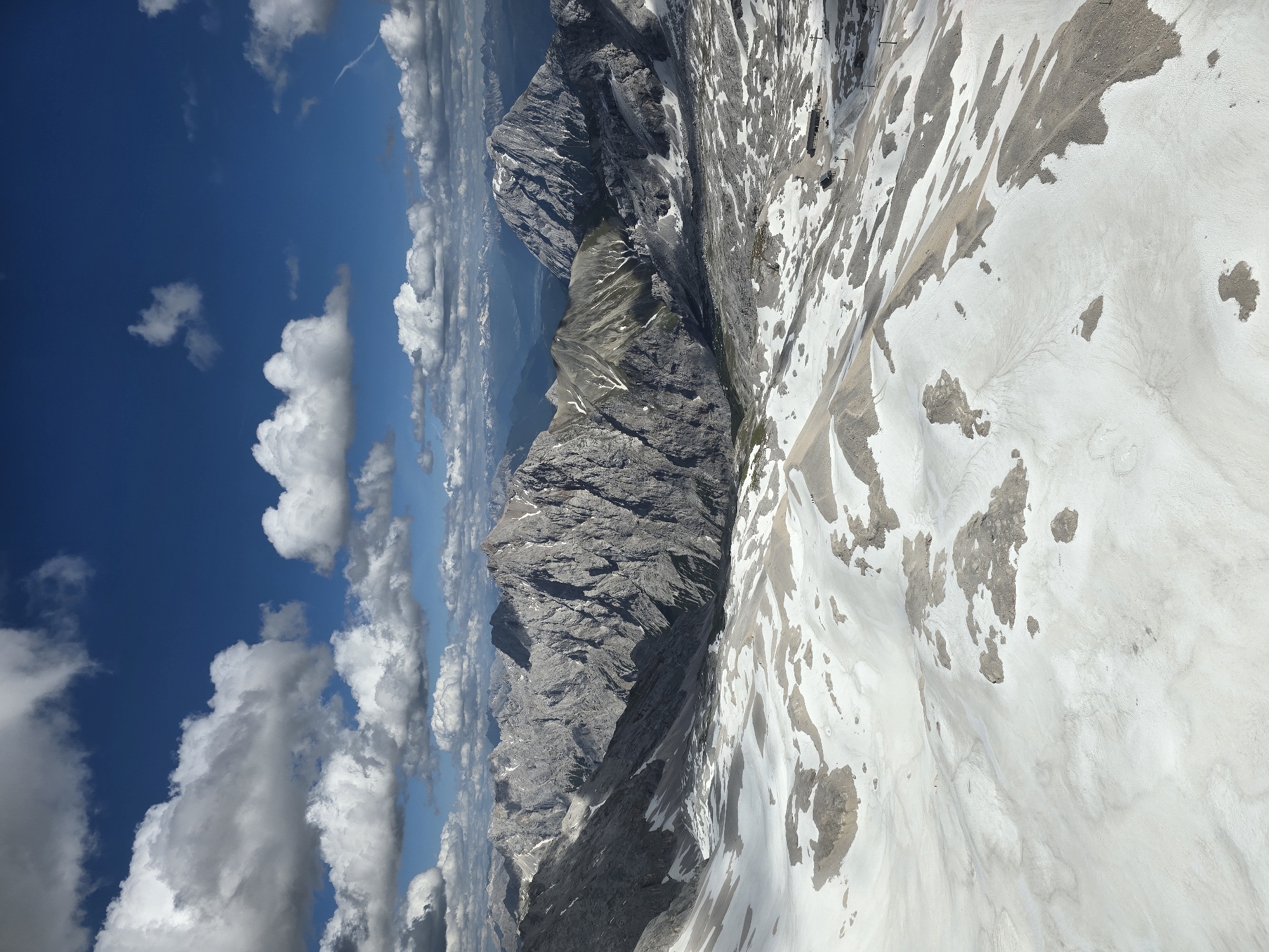 The image depicts a high-altitude mountainous landscape with a clear blue sky filled with white, fluffy clouds. The foreground is covered in snow, with patches of exposed rock and gravel. In the mid-ground and background, rugged, gray mountain peaks rise, some with snow-capped summits and rocky slopes. The scale of the mountains is vast, with layers of peaks receding into the distance. The lighting suggests a bright, sunny day.
While Garmisch-Partenkirchen is a city known for its proximity to mountains, this image specifically shows a remote, high-alpine environment. The view appears to be from a high vantage point, possibly a mountaintop or an aerial perspective, looking down over a rugged terrain that is characteristic of the Bavarian Alps. There are no urban structures or signs of civilization visible in the immediate vicinity, emphasizing the natural, wild character of the scene. A few small human figures can be observed in the distance on the snowy slopes, suggesting they are hikers or skiers. There are also some man-made structures visible in the far mid-ground, appearing to be buildings or facilities, possibly related to a ski resort or a mountain station, along with some ski lifts.