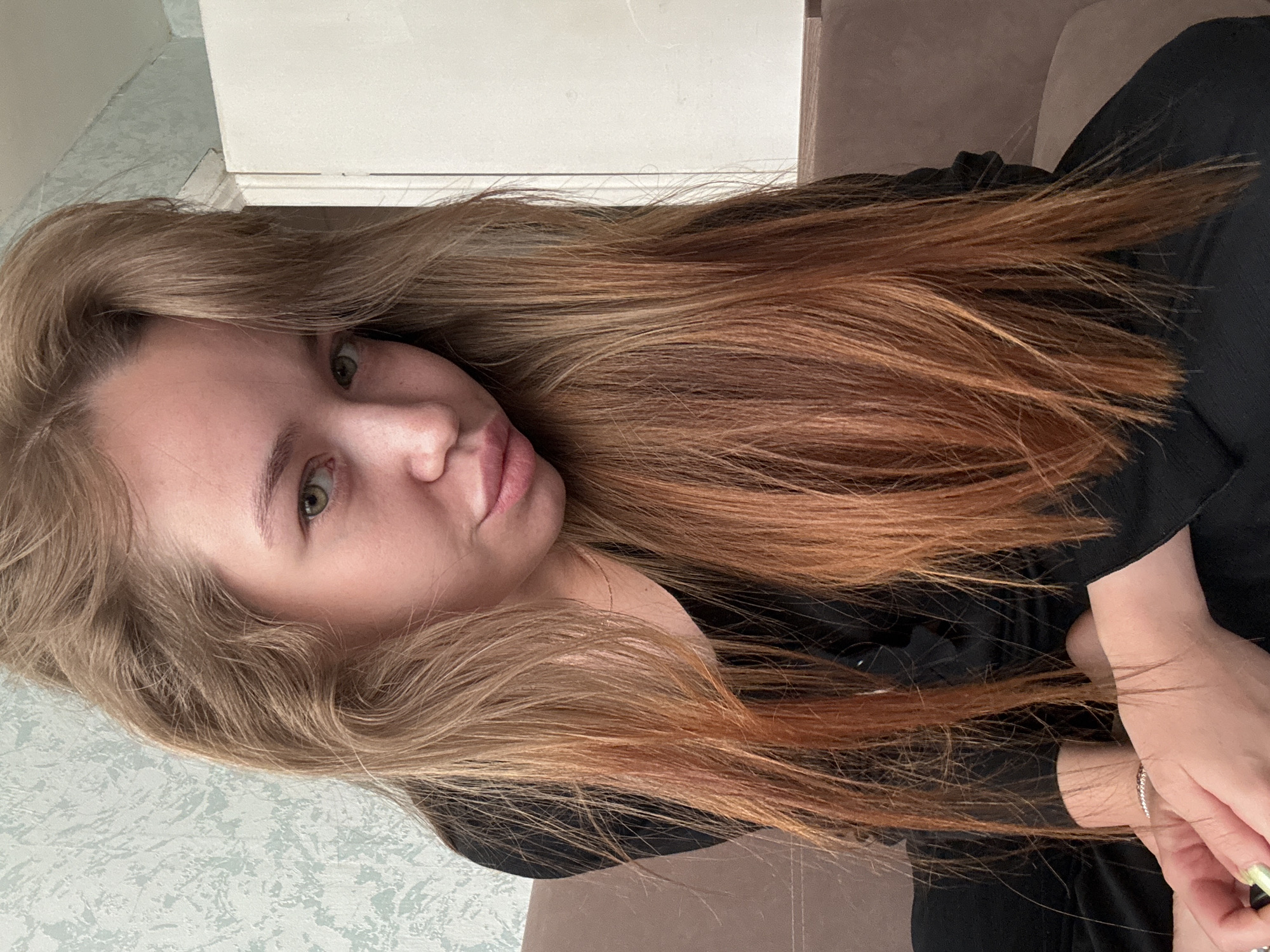 A young woman with long, blonde hair is sitting on a brown couch. She is wearing a black top. Her head is tilted to the side, and she is looking directly at the camera. The background is a wall with a light blue textured wallpaper and a white door. The scene appears to be indoors, likely in a residential setting. There are no specific city landmarks visible to identify the location as Chaikovsky, Russia.