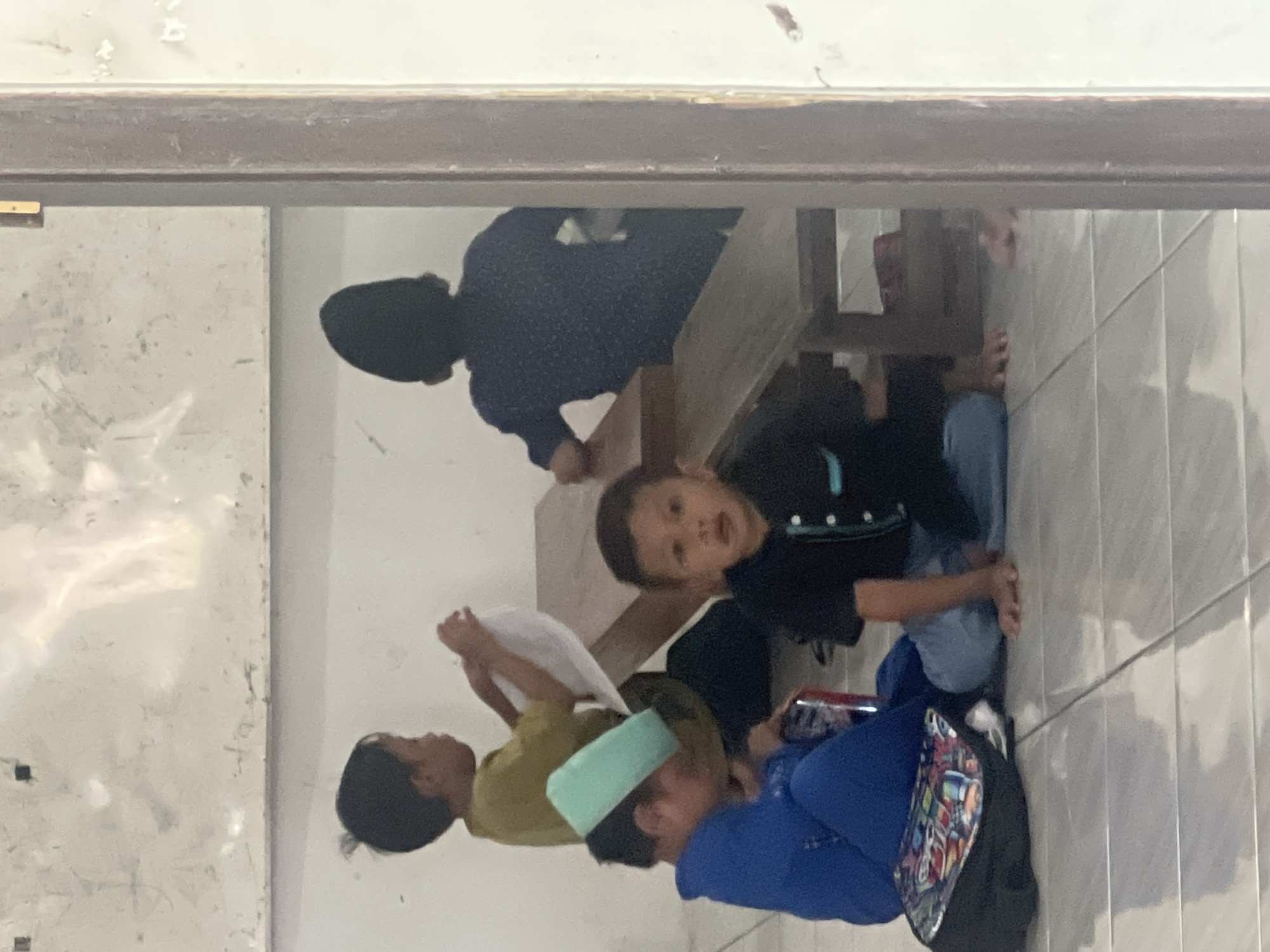 This media file shows a group of children, likely in Indonesia, given the location context of Kecamatan Pati. The image is oriented sideways, making it difficult to discern precise details about the activity.
There appear to be at least four children visible. In the foreground, a child in a blue outfit is sitting with their legs crossed, holding what looks like a cell phone or a similar electronic device. Beside them, another child in a light green hat and a yellowish shirt is seated, holding a white object that could be paper or fabric. Further back, a child in a black shirt is looking towards the camera with an expressive face. In the background, partially obscured, a person wearing a dark headscarf and a dark top is seated.
The setting appears to be indoors, possibly a classroom or a common area. The floor is tiled with light-colored square tiles. There is a wooden structure, possibly a bench or table, in the background. The walls are plain and off-white, with some visible textures or imperfections.
The children's expressions are varied. The child in black seems alert and curious, while the others are less clearly visible. There are no obvious cues about the time of day or weather conditions. The overall impression is a casual gathering of children. No visible text is present in the image.