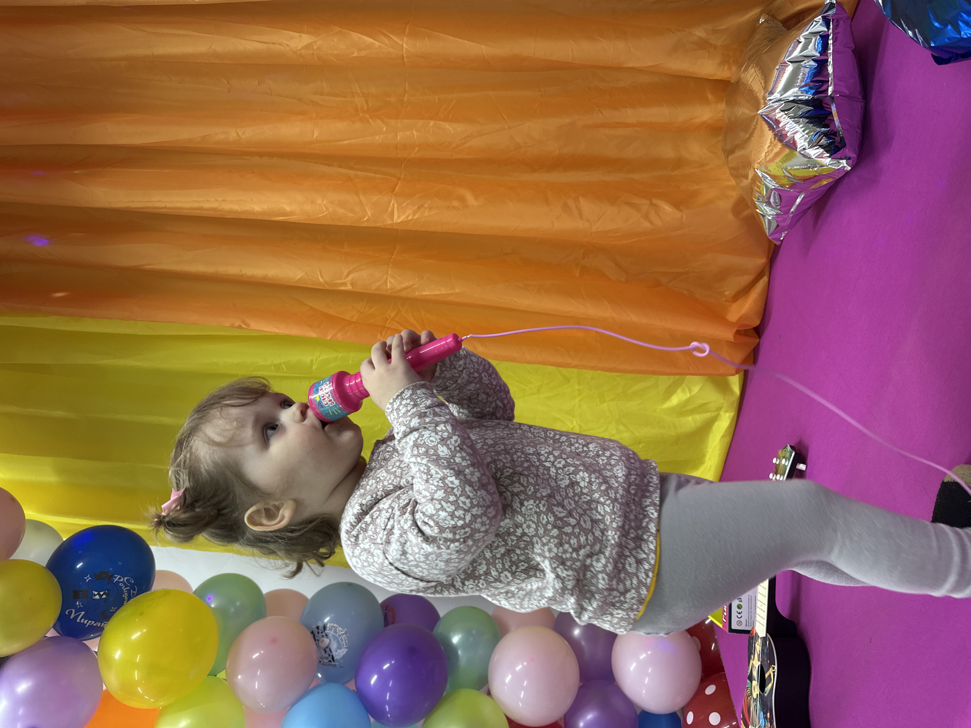 A young child is standing in front of a backdrop of orange and yellow fabric, holding a pink microphone to their mouth. The child is wearing a patterned shirt and gray leggings. Below the child are several balloons of various colors including yellow, blue, green, pink, and purple. To the right of the child, there is a purple surface with a silver balloon. A toy guitar is also visible. The location is Chaikovsky, Russia.