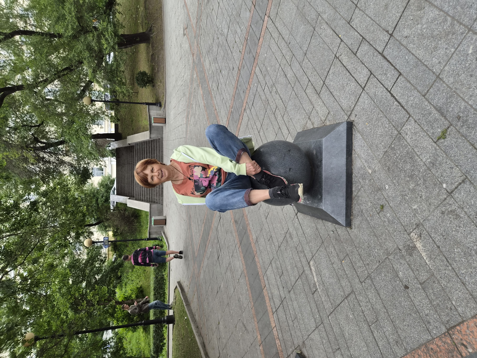 A woman with short red hair and glasses sits cross-legged on a round, dark-colored sculpture in a public park area. She is wearing blue jeans, a colorful t-shirt with a floral design, and a light green hooded jacket. Her right hand rests on her right knee, and her left foot is visible under her crossed leg. The sculpture is mounted on a dark, pyramidal base.
The park setting features a paved walkway made of gray interlocking stones with subtle orange lines marking some sections. Behind the woman, a wide concrete staircase ascends a grassy slope, flanked by trees with lush green foliage. Two men are visible in the background: one on the left is wearing a light brown jacket and jeans, looking at his phone, while another man in the middle ground wears a striped shirt and shorts, walking away from the camera. Lampposts are present in the background, and buildings are visible beyond the trees at the top of the slope. The overall environment appears to be a well-maintained urban park.