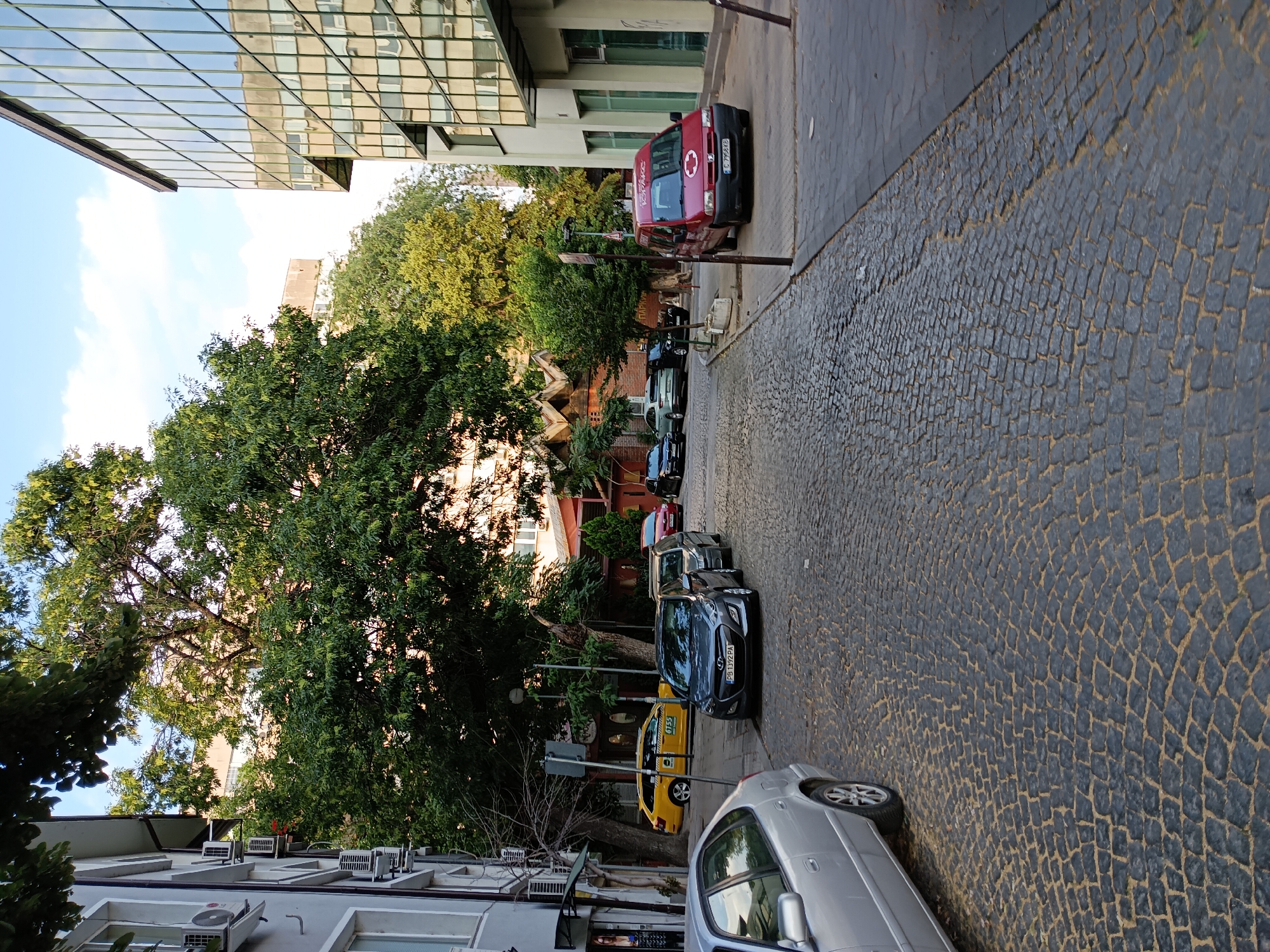 The image depicts a cobblestone street in Plovdiv, Bulgaria, likely in a mixed-use area with residential and commercial buildings. The street is lined with parked cars, including a yellow taxi, several sedans, and a red van. Lush green trees overhang the street, casting shadows on the ground. In the background, modern glass-fronted buildings stand alongside older brick structures. The sky is partly cloudy. No people are visible in the scene.