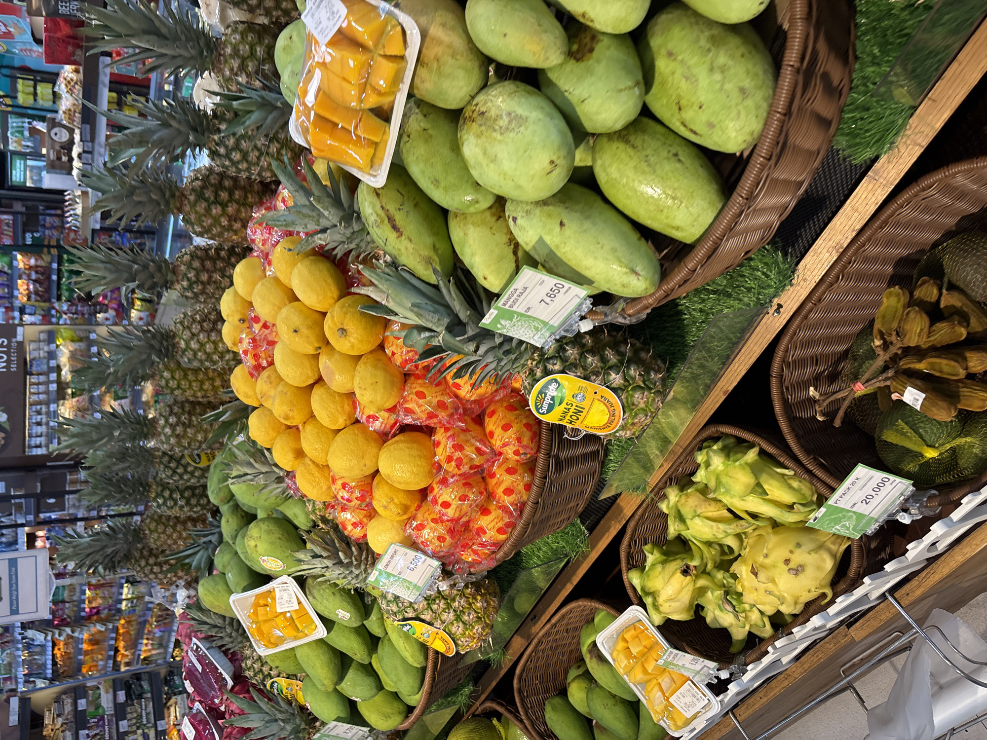 The image shows a variety of fruits displayed in wicker baskets on shelves in a store. The fruits include pineapples, lemons, mangos (some peeled and packaged), and yellow dragon fruit. The fruits have price tags indicating they are for sale. The location is likely a supermarket or fruit market in Kuta Selatan, Indonesia, based on the price tags in Indonesian Rupiah (IDR). The shelves are made of wood. The aisles of a supermarket are visible in the background.