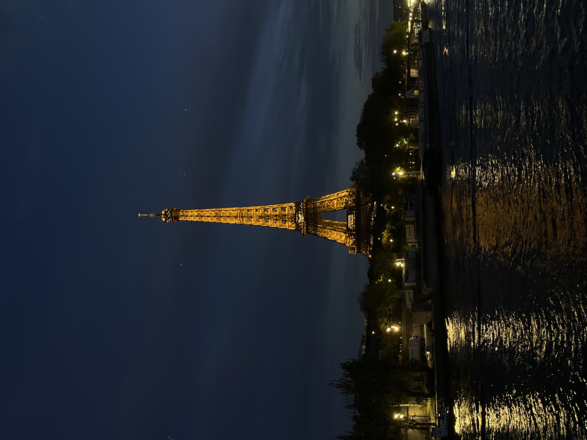 The image displays the iconic Eiffel Tower in Paris, France, illuminated at night. The photograph, originally oriented vertically but presented here rotated 90 degrees counter-clockwise, captures the monument from a vantage point across the Seine River.
The Eiffel Tower dominates the left side of the frame (when viewed upright), glowing brightly in a warm, golden light that accentuates its intricate ironwork. Its beacon is visible at the very top. The sky is a deep, dark blue, with faint, lighter cloud formations visible, suggesting a clear evening or late twilight. A few minuscule white specks are scattered across the dark expanse of the sky.
To the right of the Eiffel Tower, the dark waters of the Seine River flow, reflecting the city's artificial lights and the tower's golden glow. These reflections appear as shimmering, elongated streaks of light dancing on the water's rippled surface. Along the riverbank, silhouetted trees and various structures are visible, punctuated by numerous bright streetlights and building lights. These urban lights contribute to the ambient glow and the reflections on the river.
The scene conveys a peaceful nighttime atmosphere, focusing on the architectural grandeur of the Eiffel Tower and its integration with the Parisian landscape by the river. There are no visible people or specific activities taking place, and no discernible text is present within the image. The time of day is clearly night, characterized by the artificial illumination and dark sky.