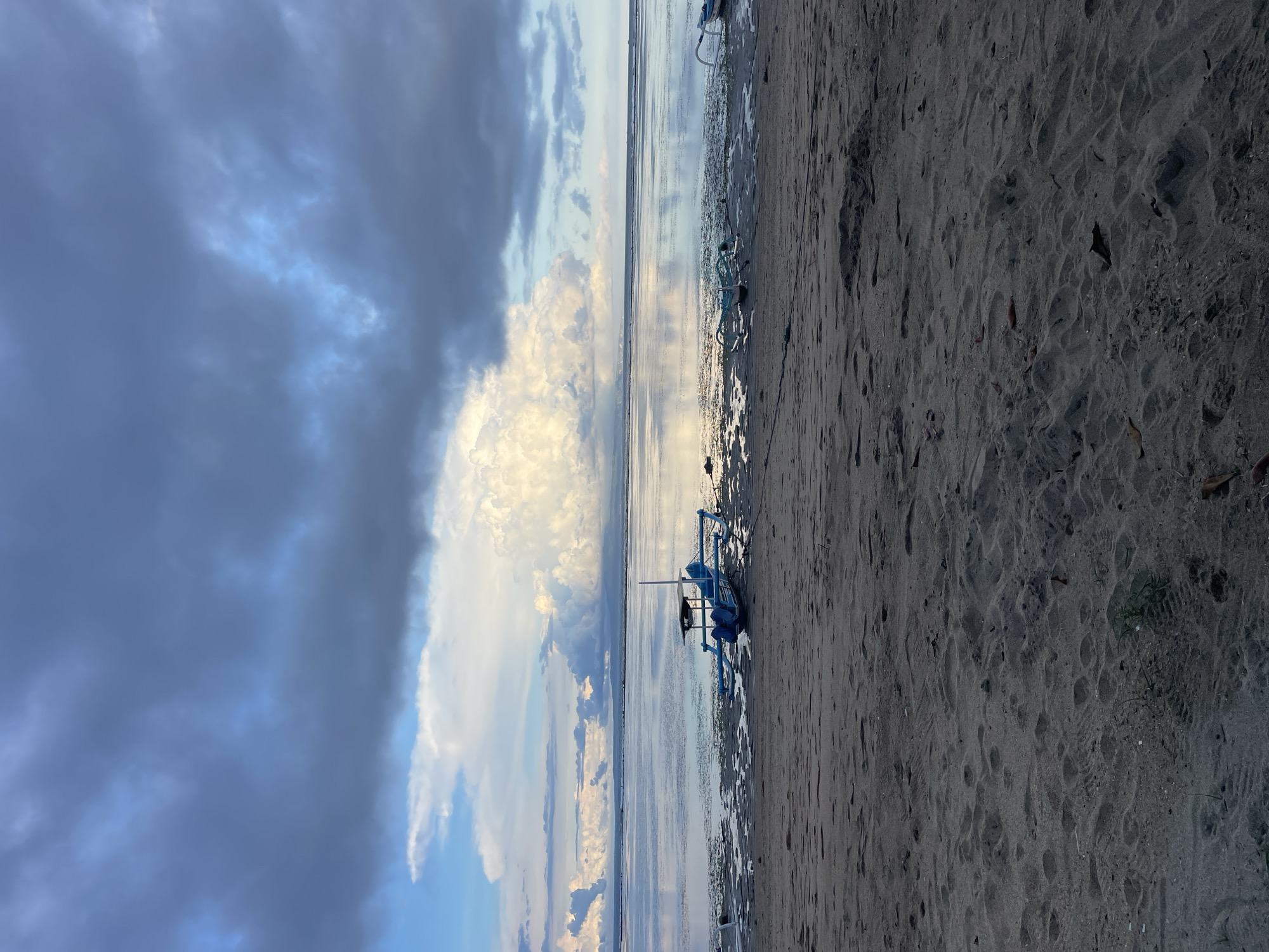 The image depicts a beach scene in Denpasar, Indonesia. The sky is overcast with a mix of dark storm clouds and a large, bright white cumulus cloud formation near the horizon. A calm body of water, possibly the ocean or a bay, reflects the sky's colors. On the sandy shore, a blue, outrigger-style fishing boat rests. The sand is damp and dark, with some debris scattered along the shoreline. No people are visible in the frame.